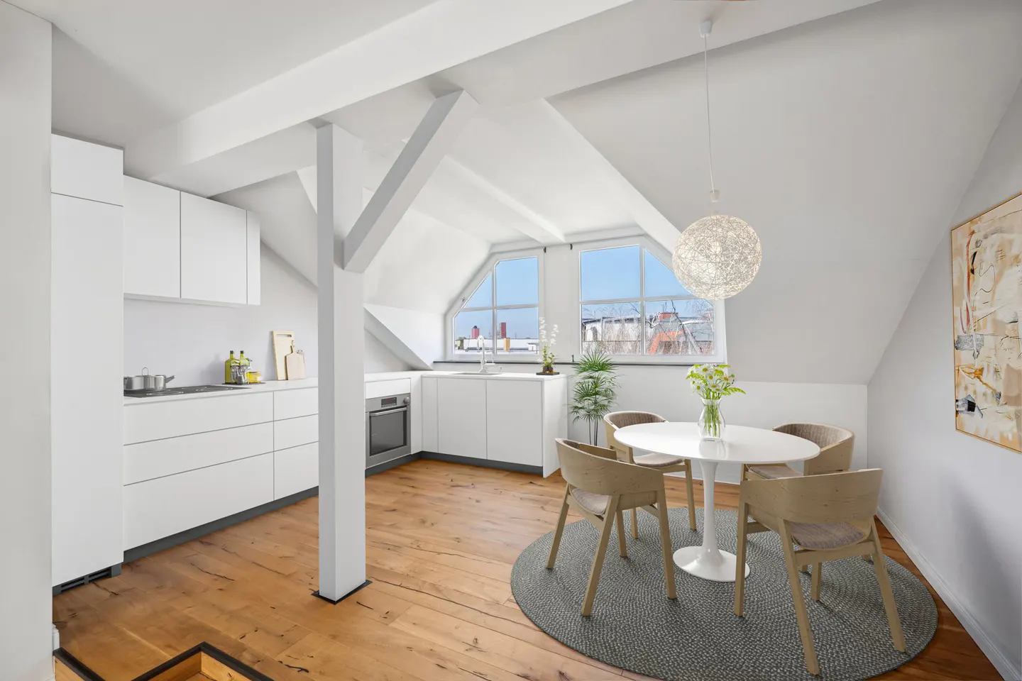 Bright, modern attic apartment with white kitchen, wood floors, and dining area with round table and four chairs. Windows overlook city.