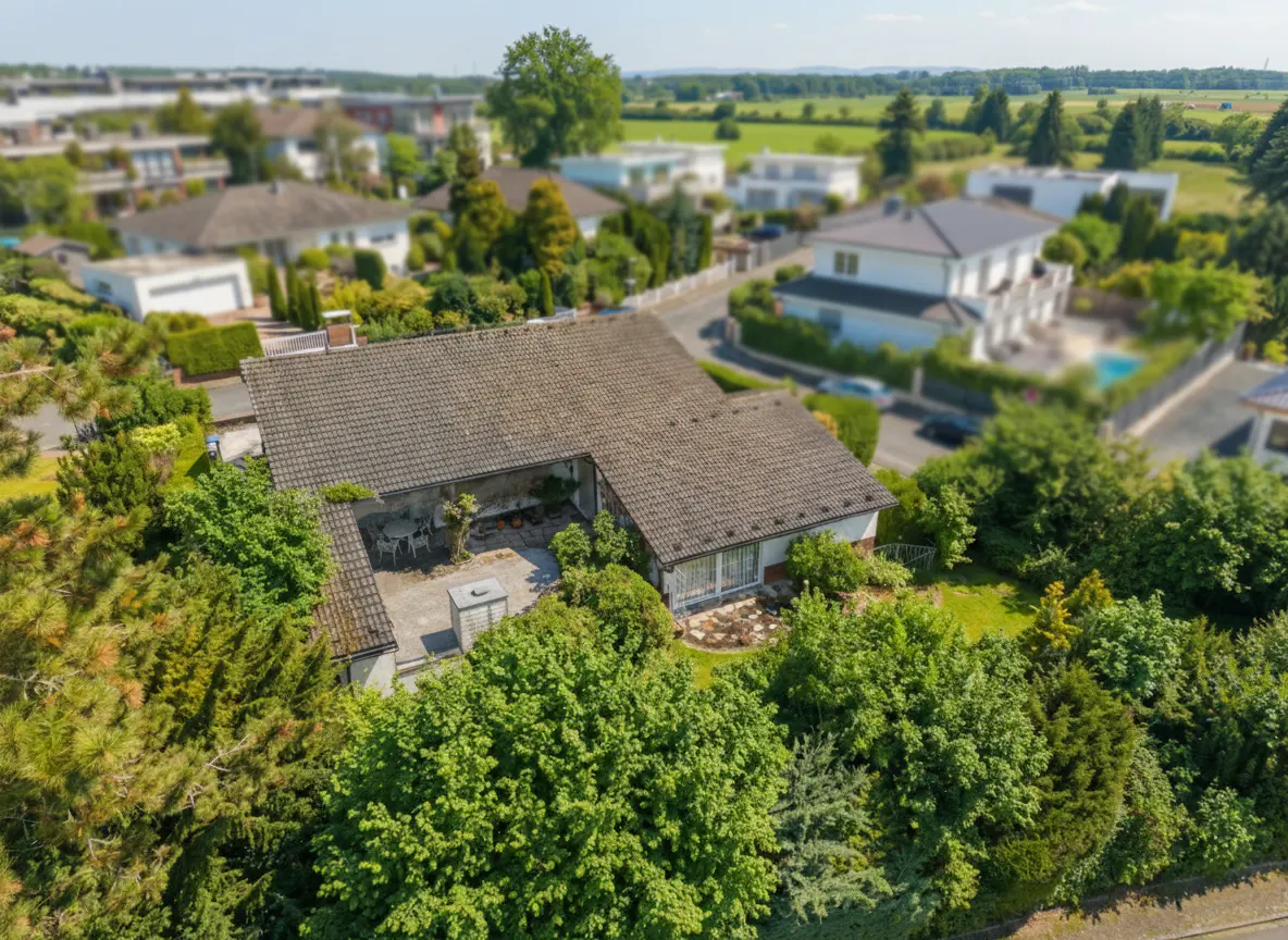 Aerial view of a single-story house with a gray tiled roof, surrounded by lush green trees and other houses in the background.
