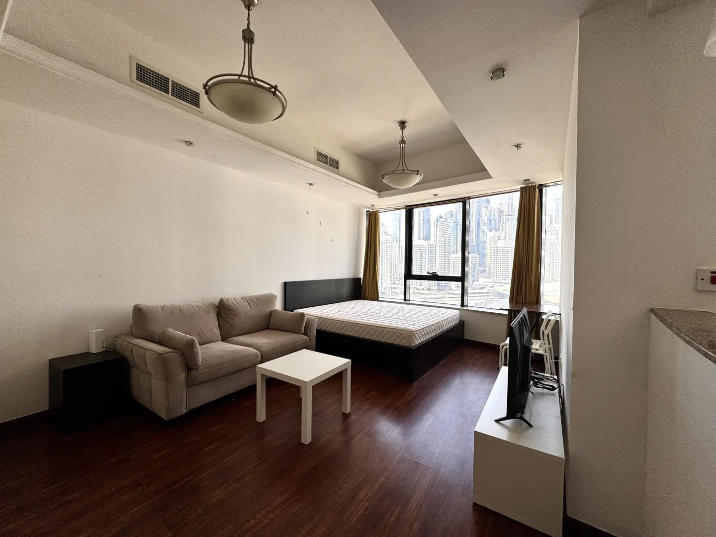 Studio apartment with a beige sofa, white table, bed, and TV. Large window with city view. Brown wood floors.