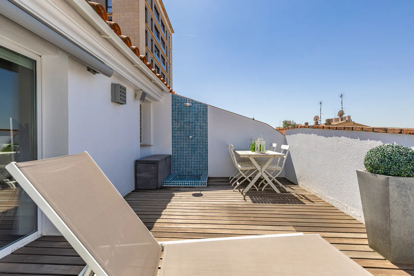 Rooftop patio with wooden deck, white walls, and blue tiled shower. A lounge chair, table, and potted plant add to the outdoor space.