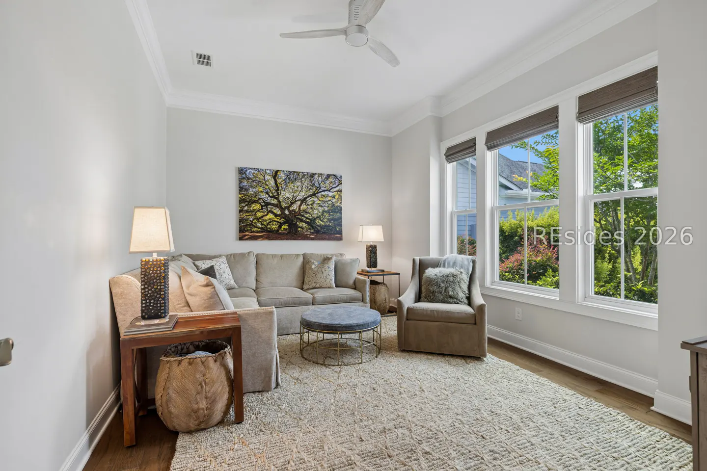 Bright living room with a beige sectional, armchair, round ottoman, and large windows overlooking a green garden.