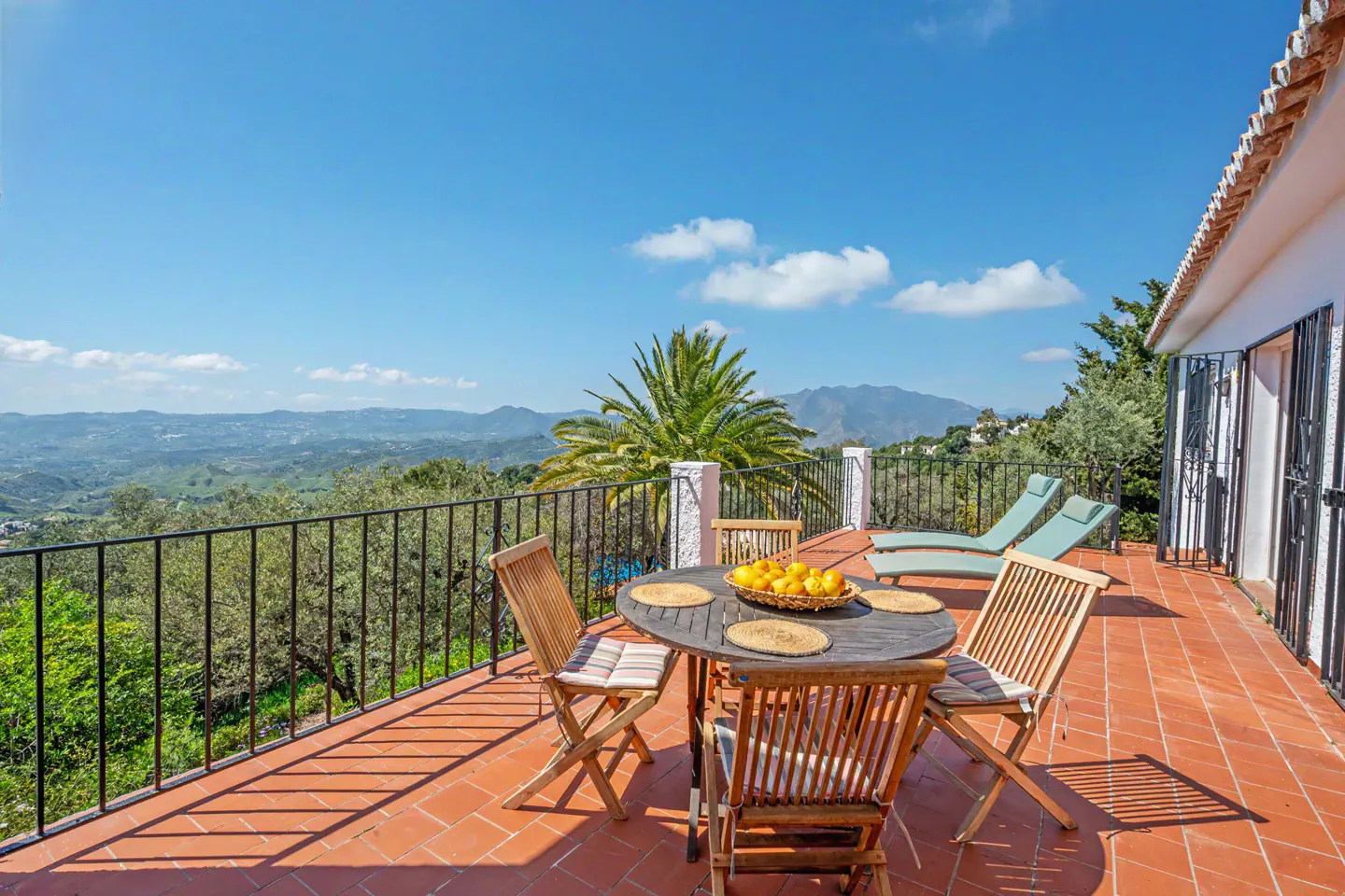 Terrace view with table, chairs, and lounge chairs overlooking mountains. Blue sky.