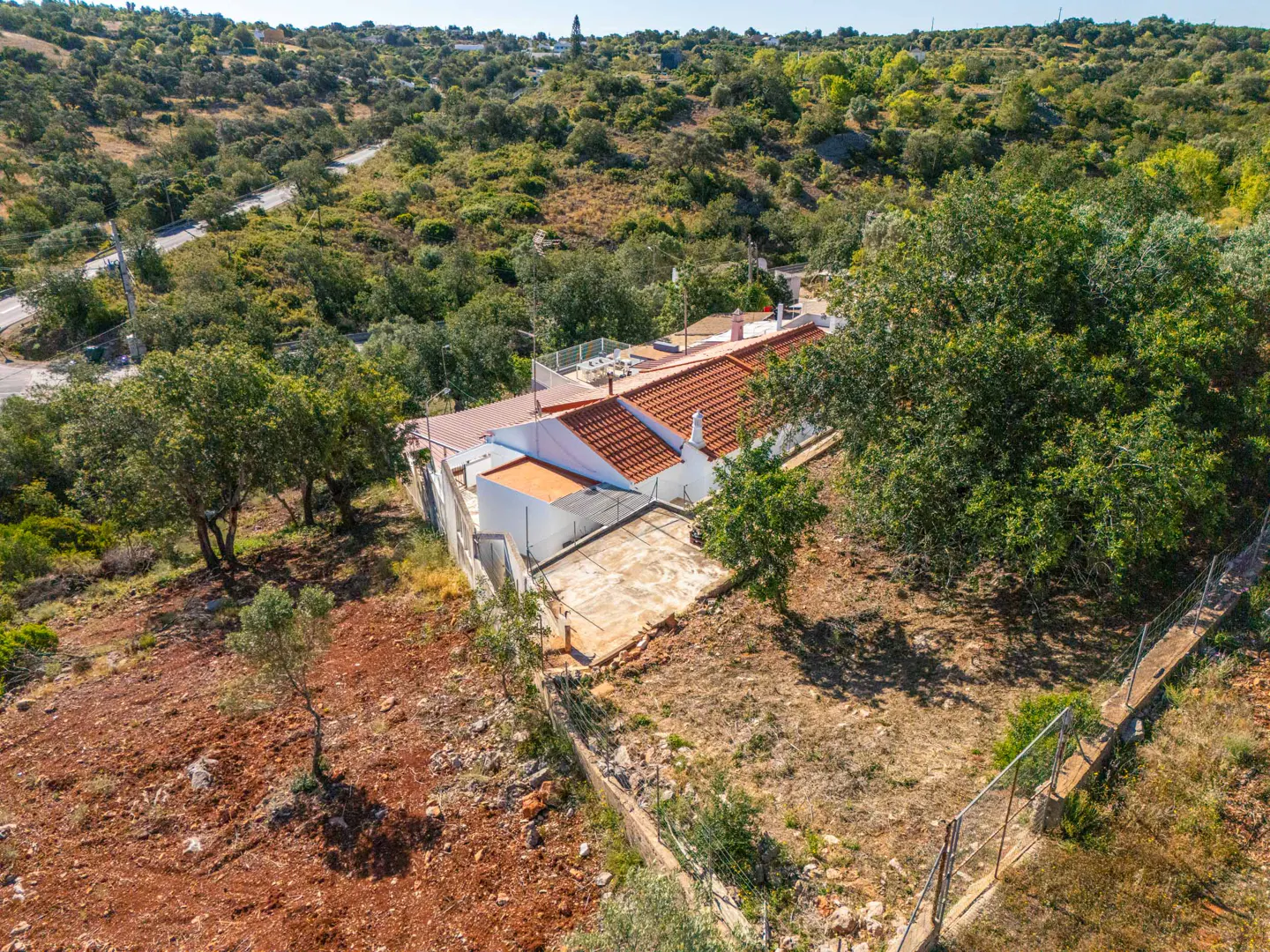 Aerial view of a white house with a red tile roof, surrounded by green trees and a hillside. A road winds through the trees in the background.