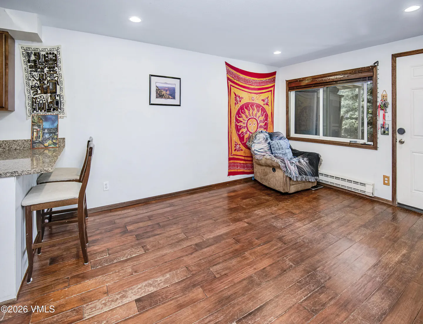 A room with hardwood floors, a breakfast bar with two stools, a recliner under a sun tapestry, and a window.