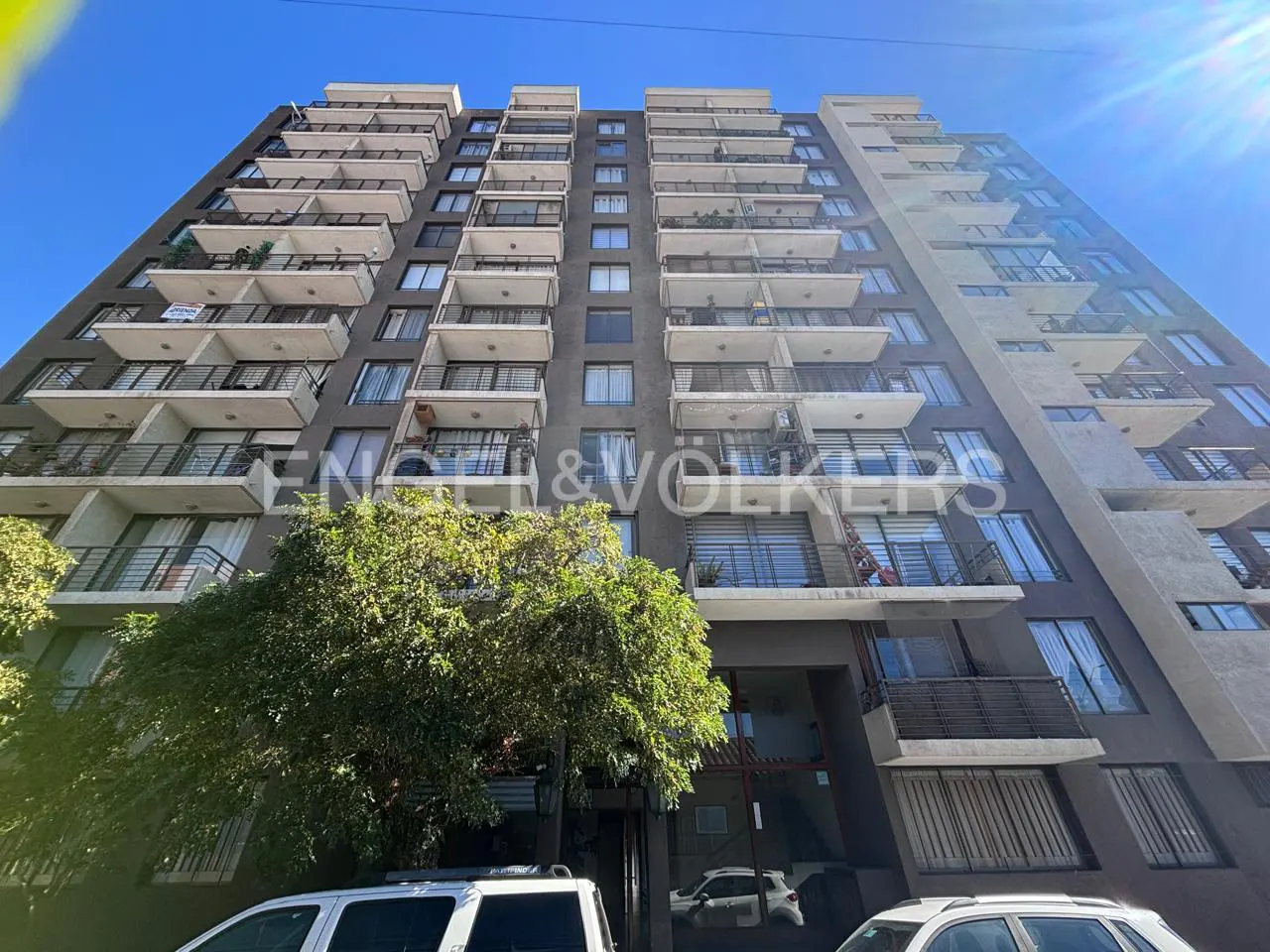 Exterior view of a tall, gray apartment building with white balconies under a blue sky. Engel & Völkers logo is visible.