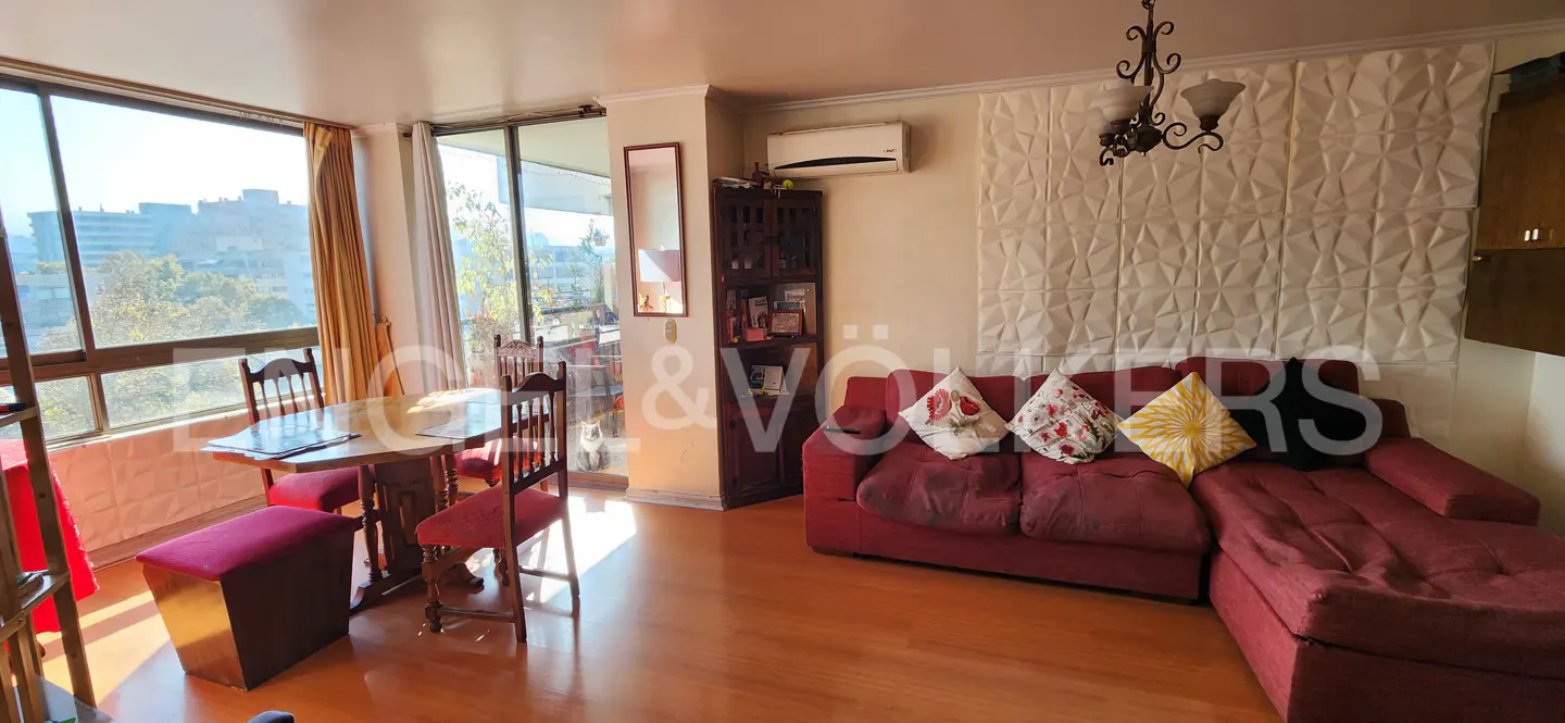 Bright living room with wood floors, red sofa with pillows, dining table, and large windows with city views.
