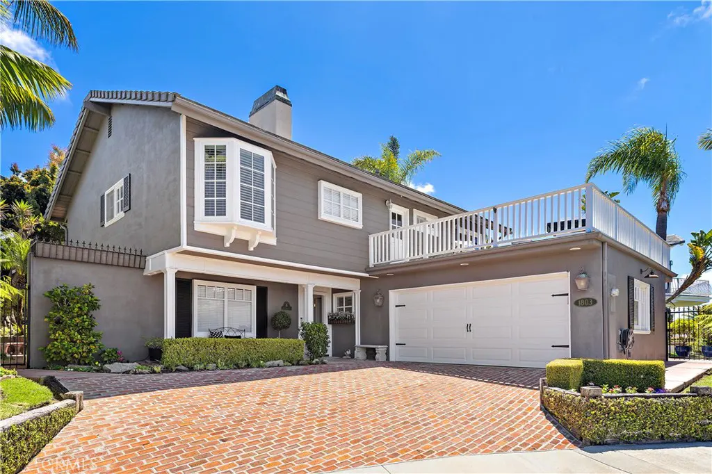Two-story gray house with a white garage, brick driveway, and second-story balcony under a clear blue sky.