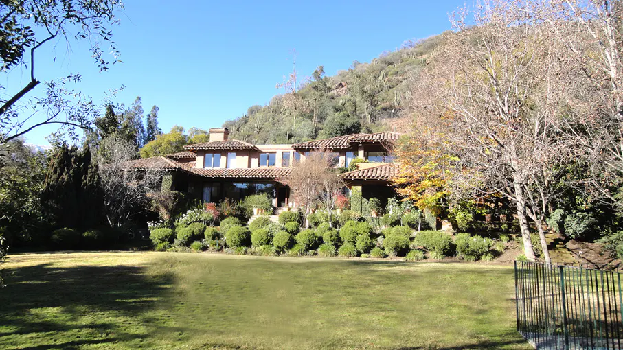 A large, tan house with a red tile roof sits on a green lawn with trees and a hill in the background.