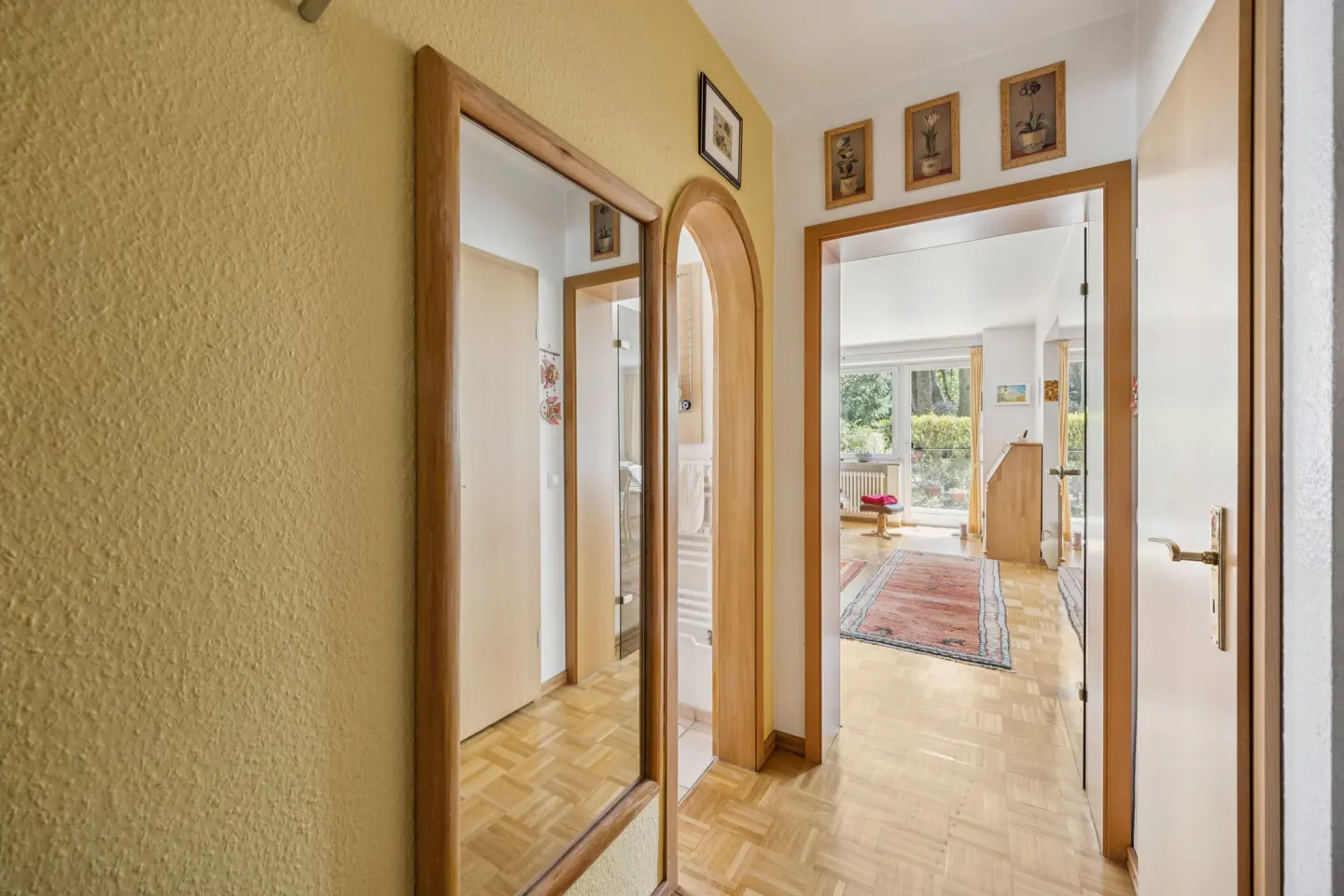 Hallway with parquet floors, yellow walls, and wooden door frames. A mirror reflects a door, and an open doorway leads to a bright living room.