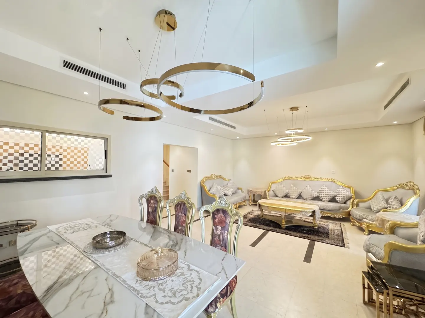 A bright, open-concept living and dining area with gold chandeliers, ornate furniture, and a marble dining table.