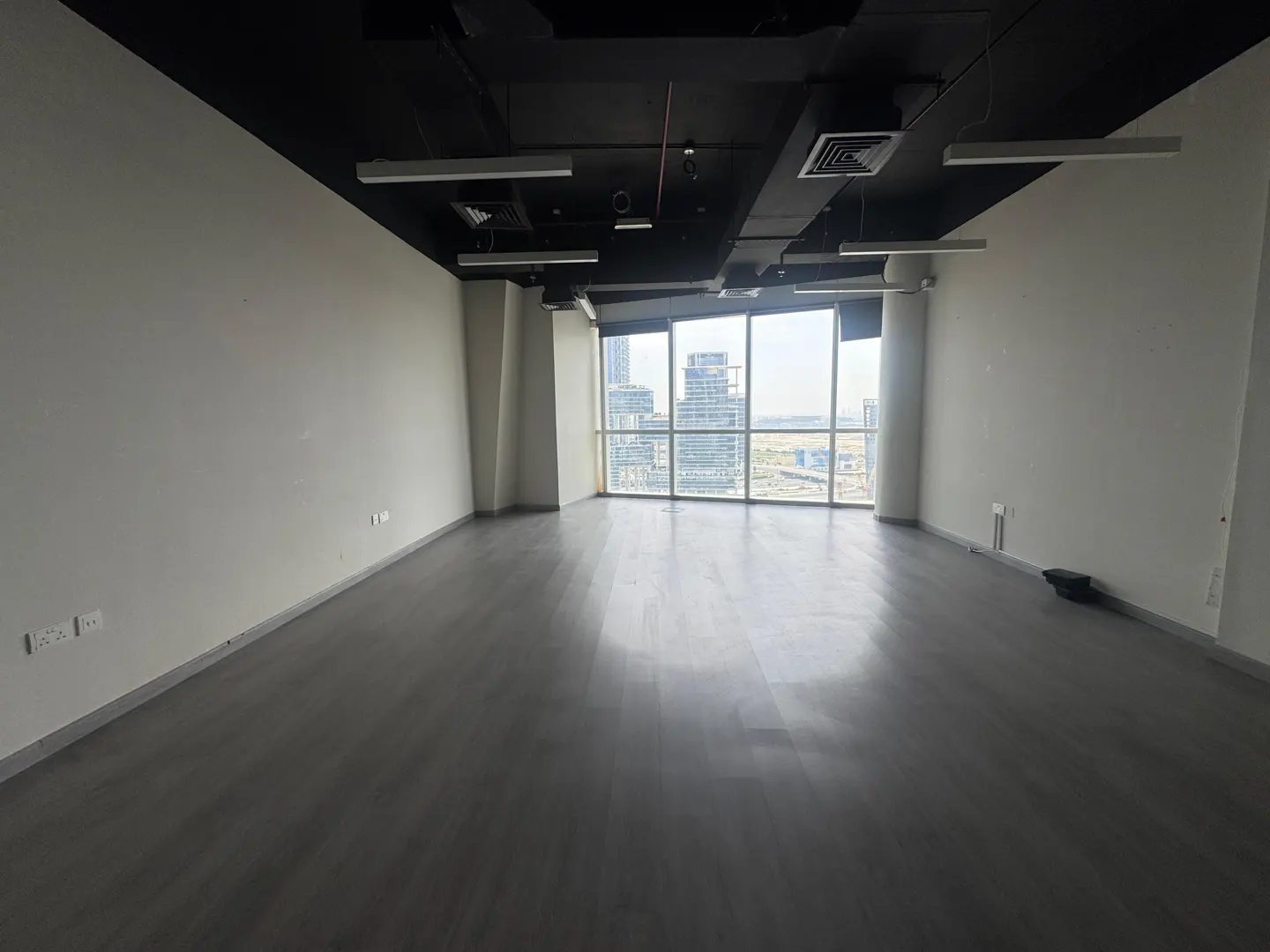Empty office space with gray floors, white walls, and a black ceiling. Large windows offer a city view.