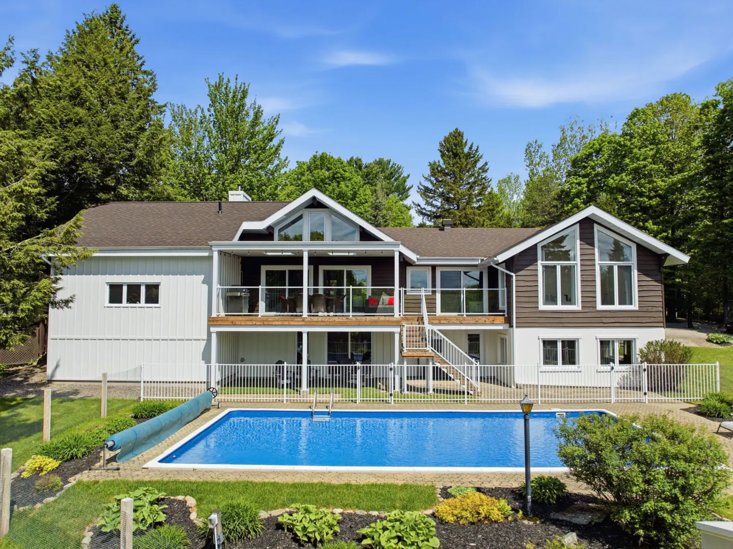 Two-story house with a pool. The house is white and brown with a deck and large windows. A white fence surrounds the pool.