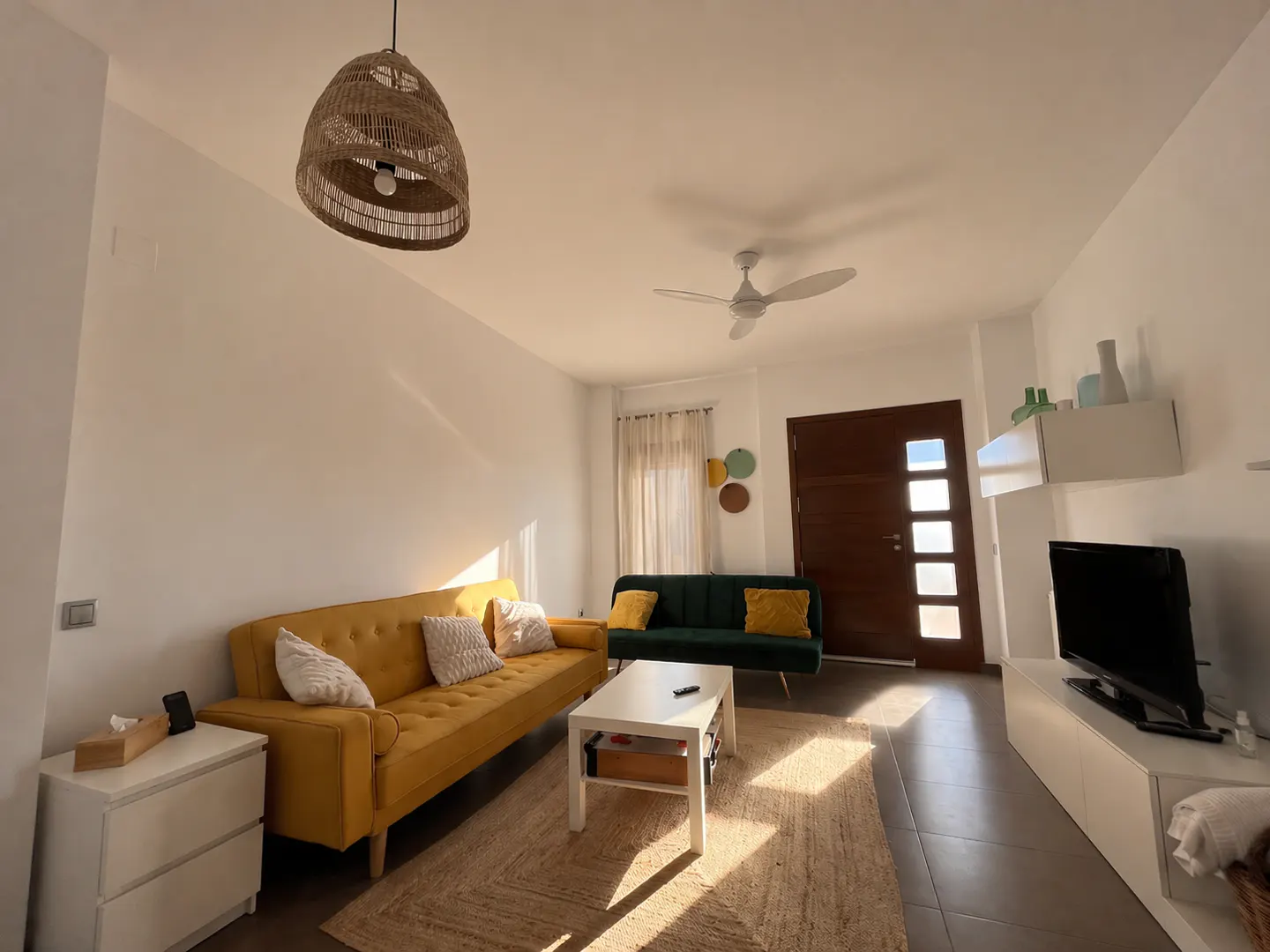 A bright living room with a yellow sofa, green loveseat, and a white coffee table on a jute rug. A brown door and TV stand are visible.