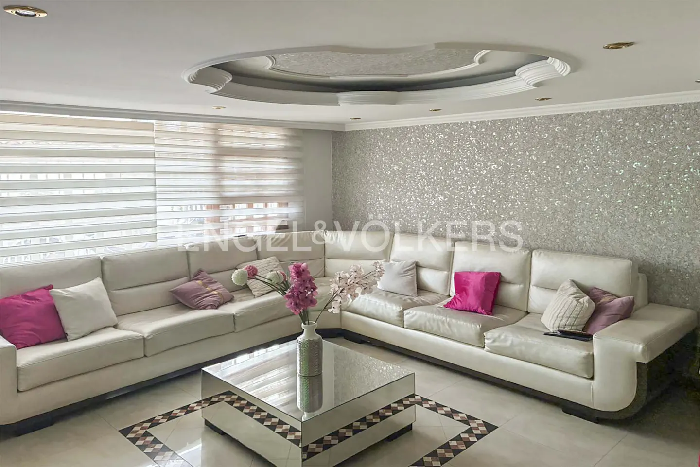 Modern living room with a large white sectional sofa, pink accent pillows, and a mirrored coffee table against a glitter wall.