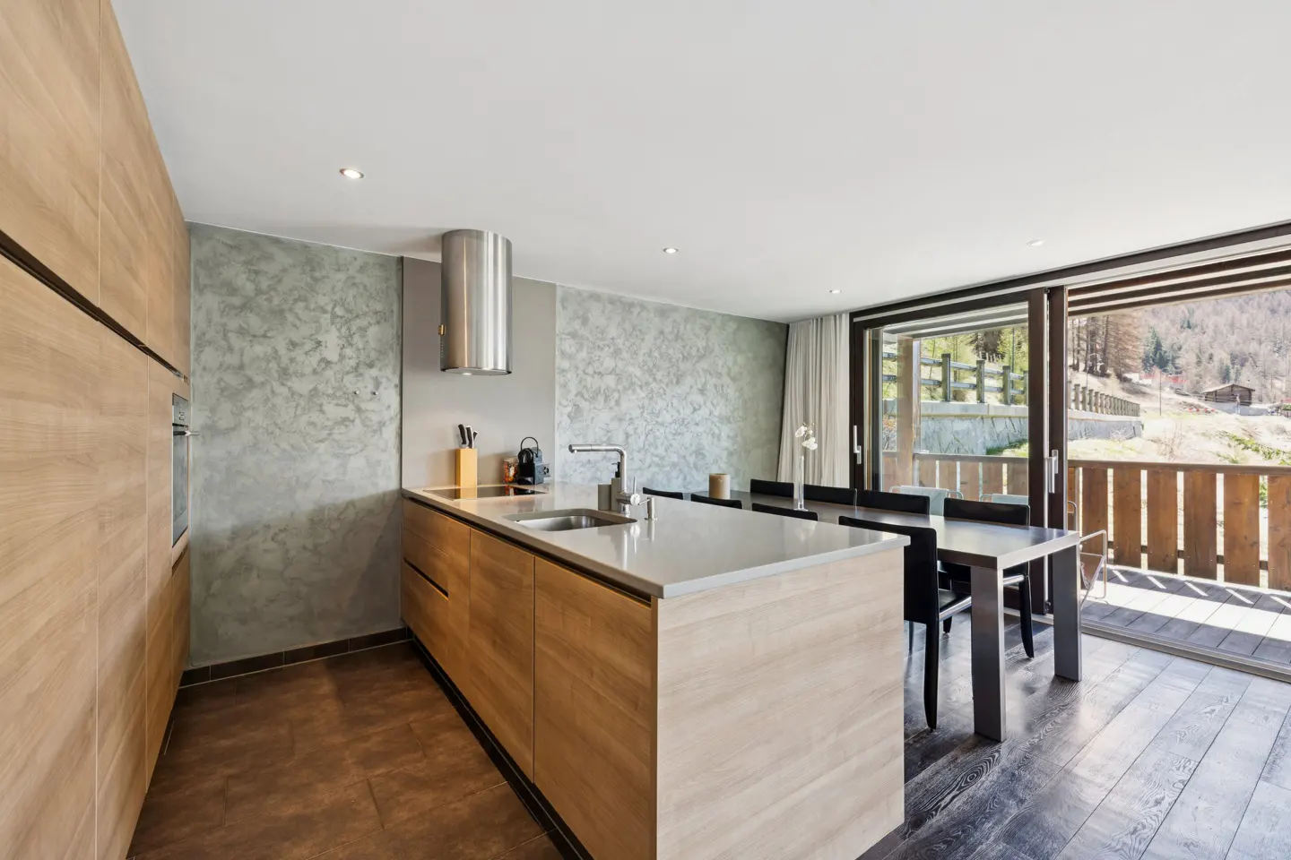 Modern kitchen with light wood cabinets, stainless steel hood, and island. Dining table and balcony with mountain view visible through sliding glass doors.