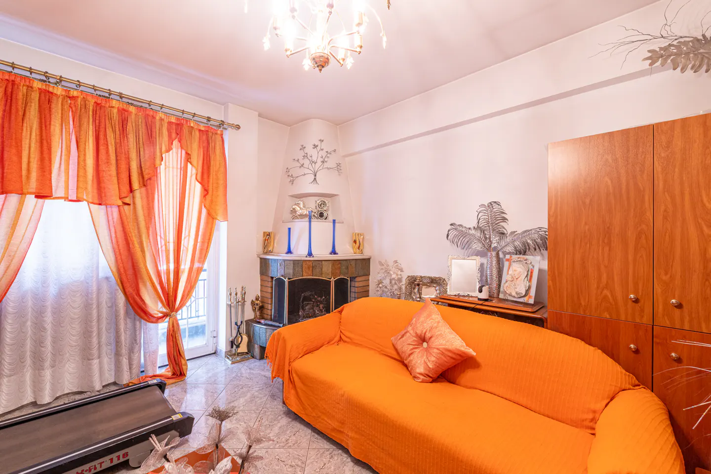 Living room with orange sofa, curtains, and wood cabinet. Fireplace with blue vases and a treadmill are also visible.