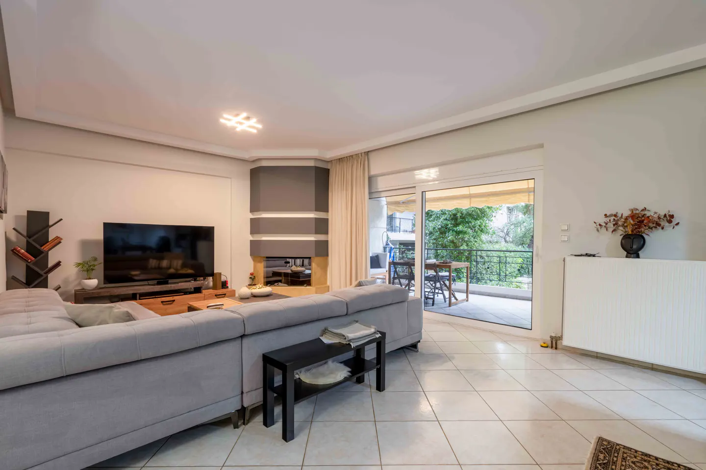 Bright living room with gray sectional sofa, TV, fireplace, and balcony with outdoor furniture. Tiled floor and neutral walls.