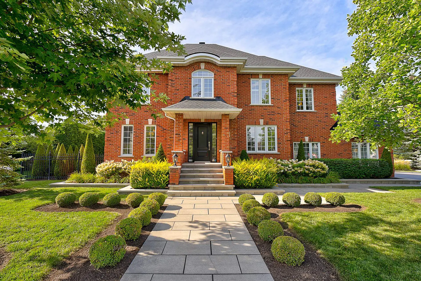 Two-story red brick house with a black front door, gray roof, and a stone walkway lined with round bushes.