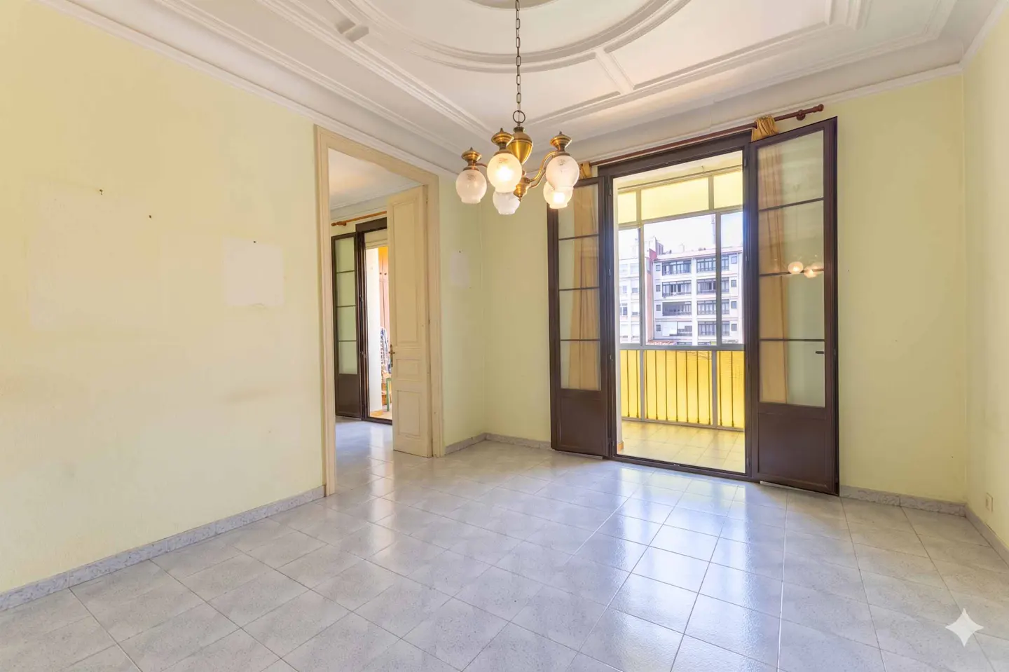 Empty room with pale yellow walls, gray tile floor, and a chandelier. Balcony doors open to a city view.