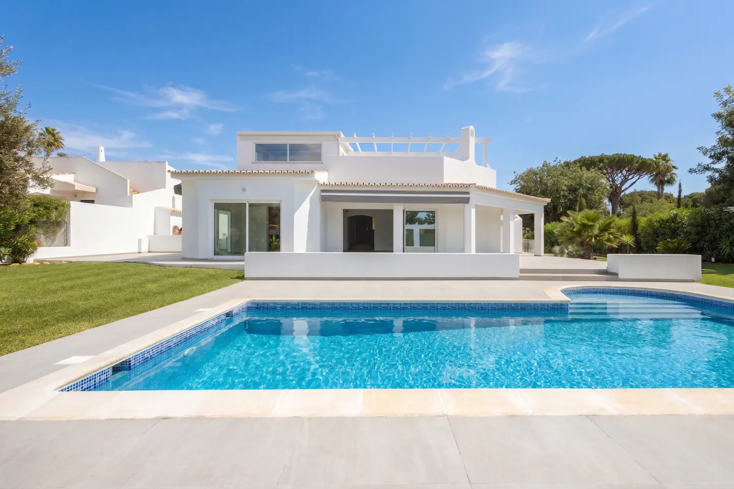 Modern white villa with a blue swimming pool, green lawn, and clear blue sky on a sunny day.