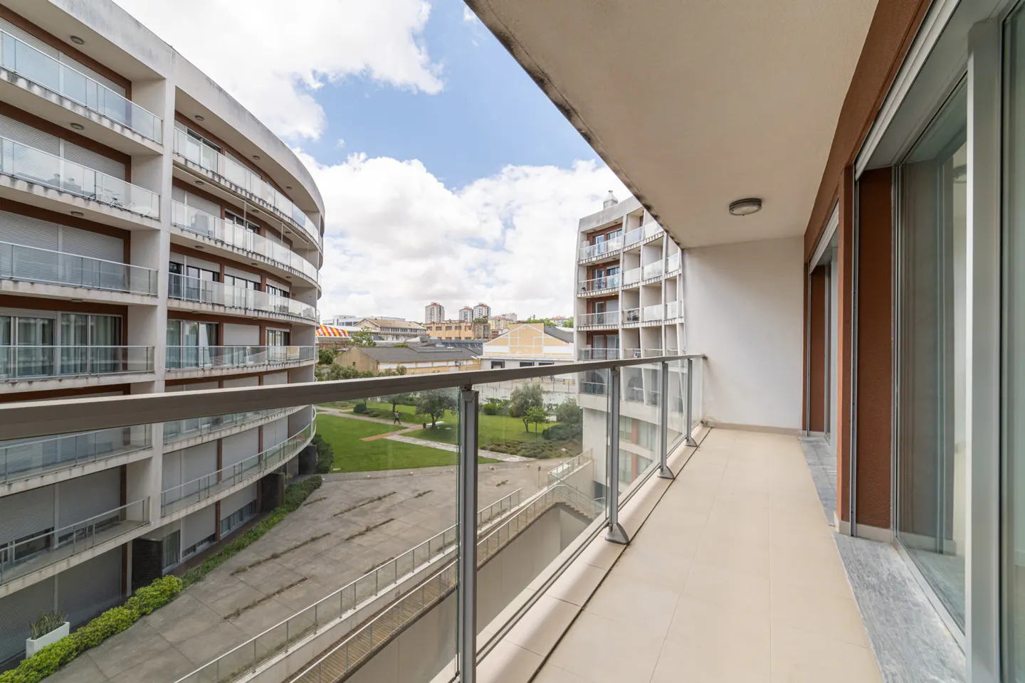 Balcony view of a modern apartment complex. Glass railings overlook a green courtyard and other buildings under a blue sky.