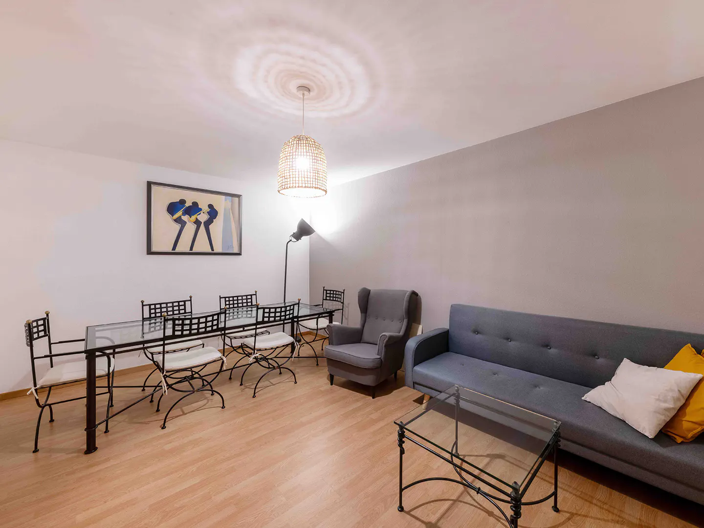 Living room with a glass dining table, gray sofa, armchair, and wood floors. A woven pendant light hangs from the ceiling.
