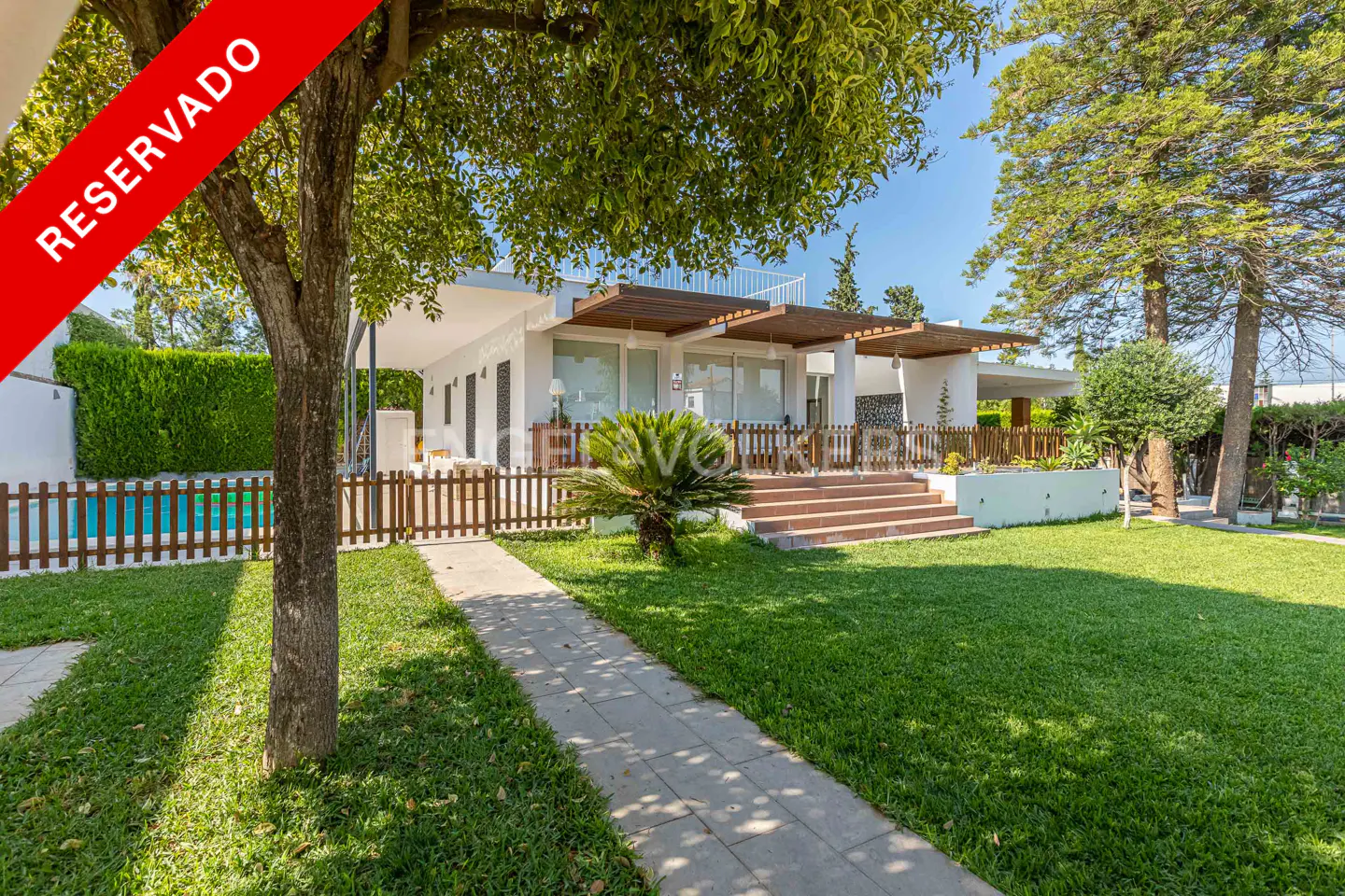 Exterior view of a white house with a green lawn, trees, and a pool. A red "Reservado" banner is in the upper left corner.