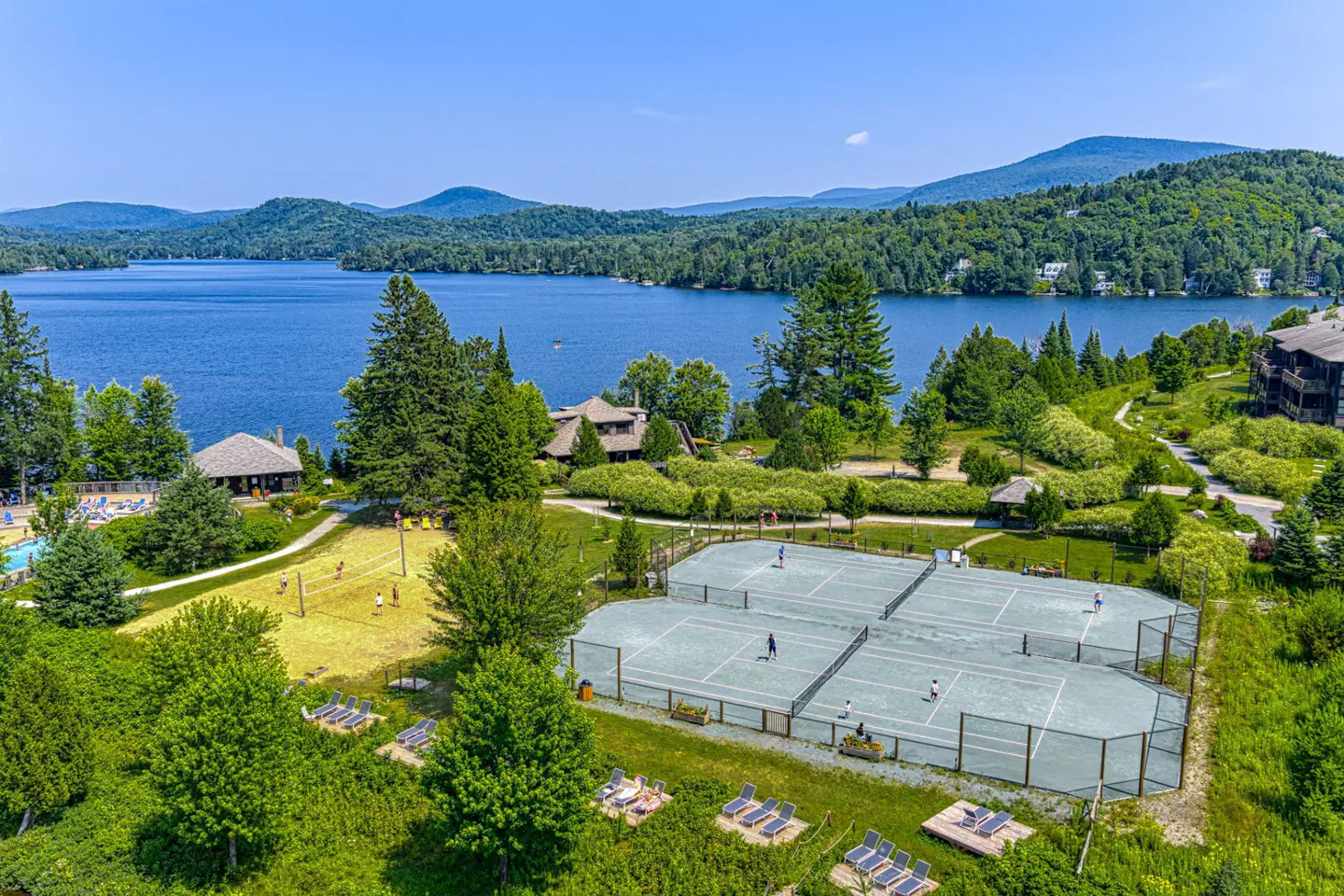 Aerial view of a resort with tennis courts, a lake, and mountains in the background on a sunny day.