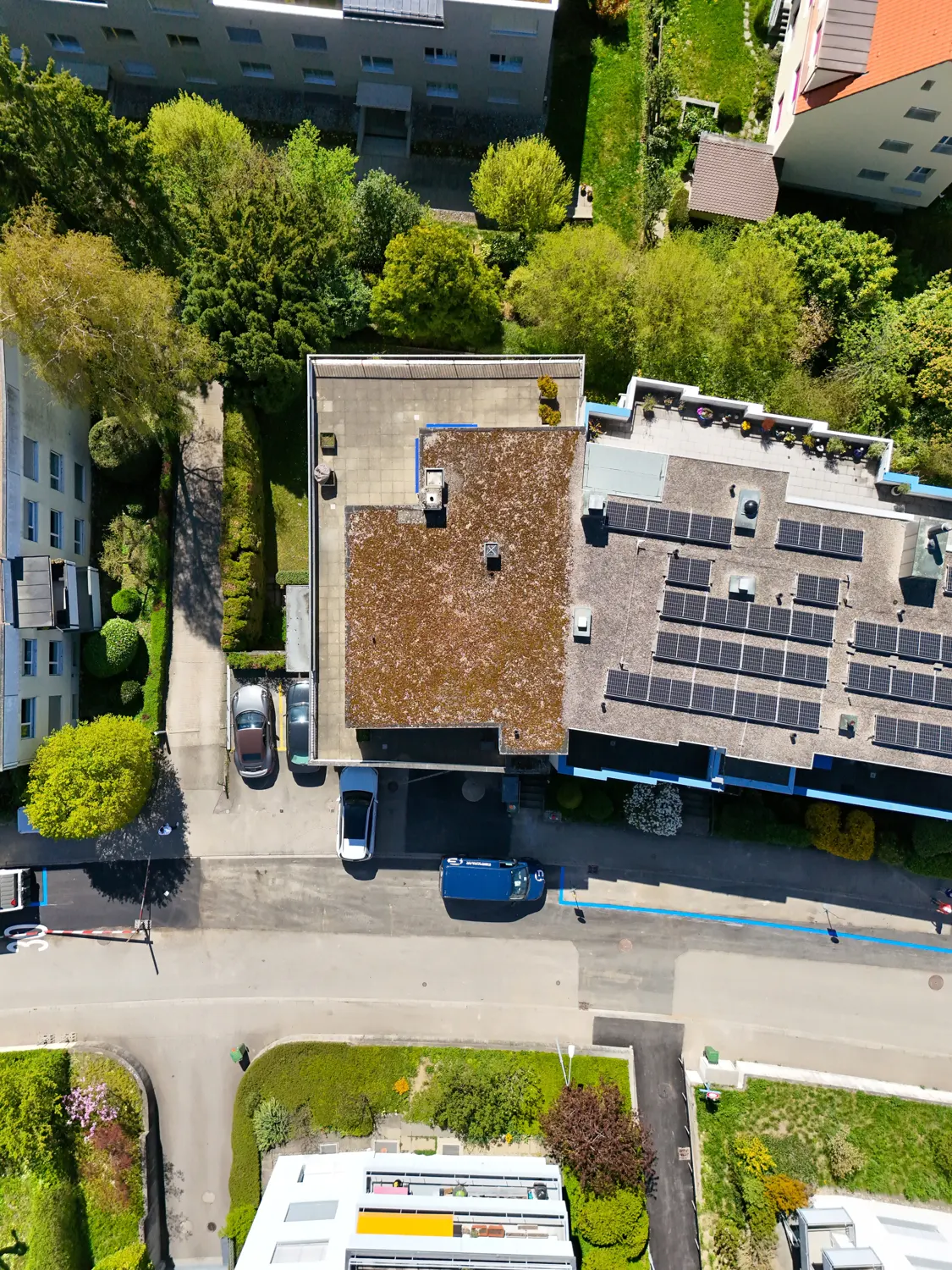 Aerial view of a building with solar panels on the roof, surrounded by green trees and other buildings. Cars are parked on the street.