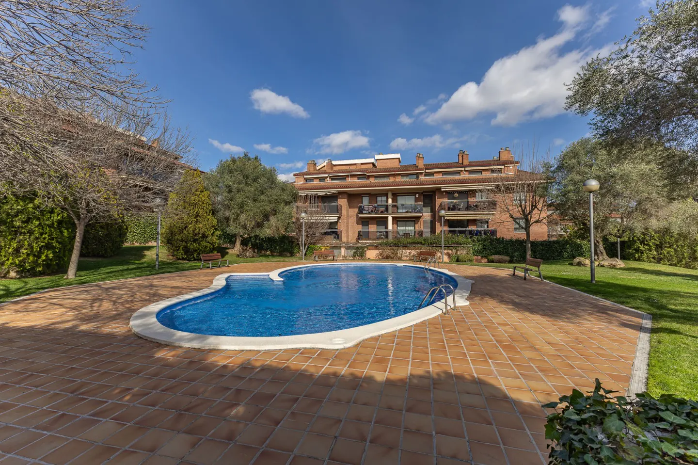 Outdoor pool with blue water and a brick patio, surrounded by green grass, trees, and a brick apartment building under a blue sky.