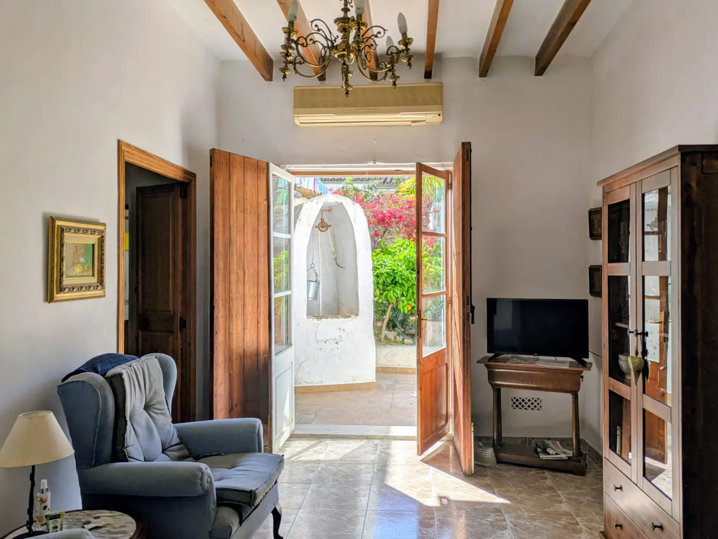 A bright living room with open wooden doors leading to a courtyard with a well and colorful plants.