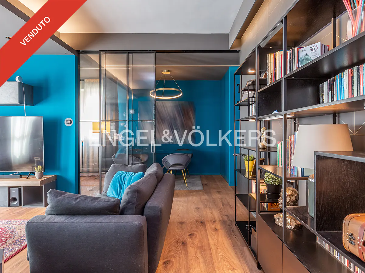 A modern living room with blue walls, a gray sofa with a blue pillow, and a black bookshelf. "Venduto" banner in the upper left corner.