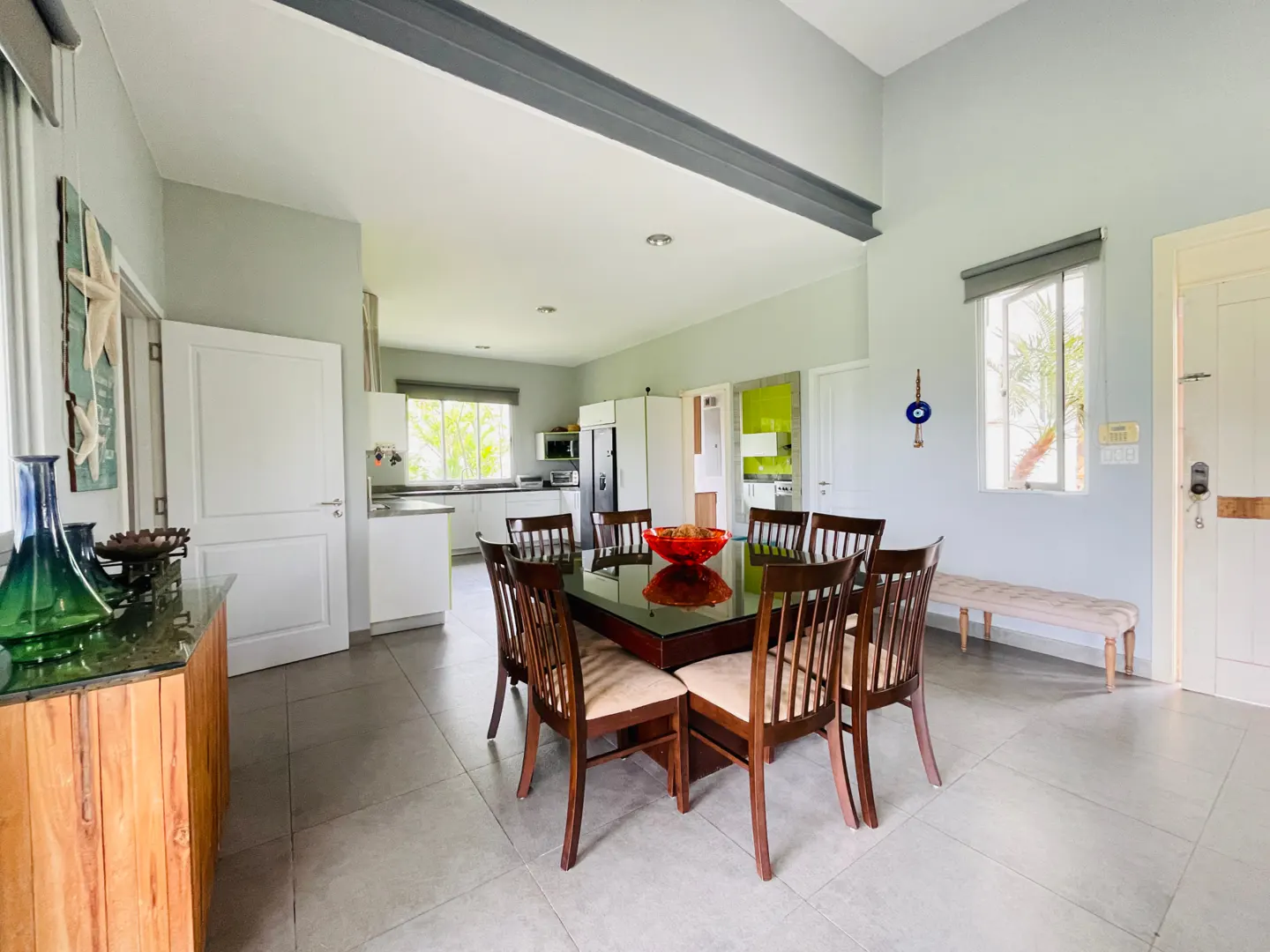 Open-concept dining room with a dark wood table, eight chairs, and a red bowl centerpiece. Kitchen in background, gray walls, and tile floor.