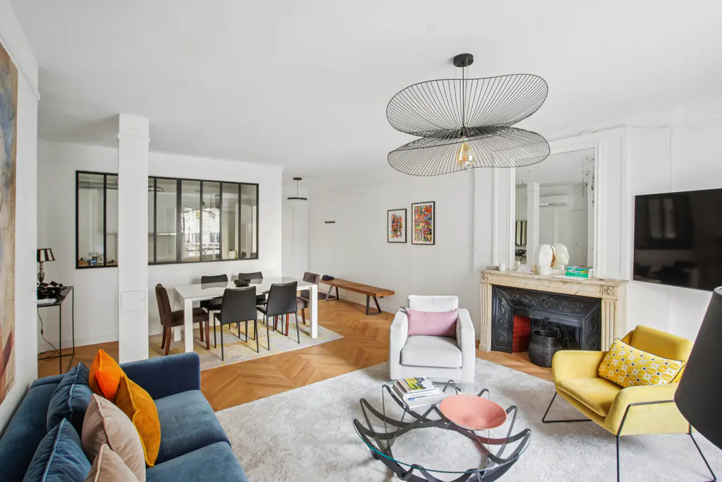 Bright living room with blue sofa, yellow chair, fireplace, and dining table. A modern chandelier hangs from the white ceiling.