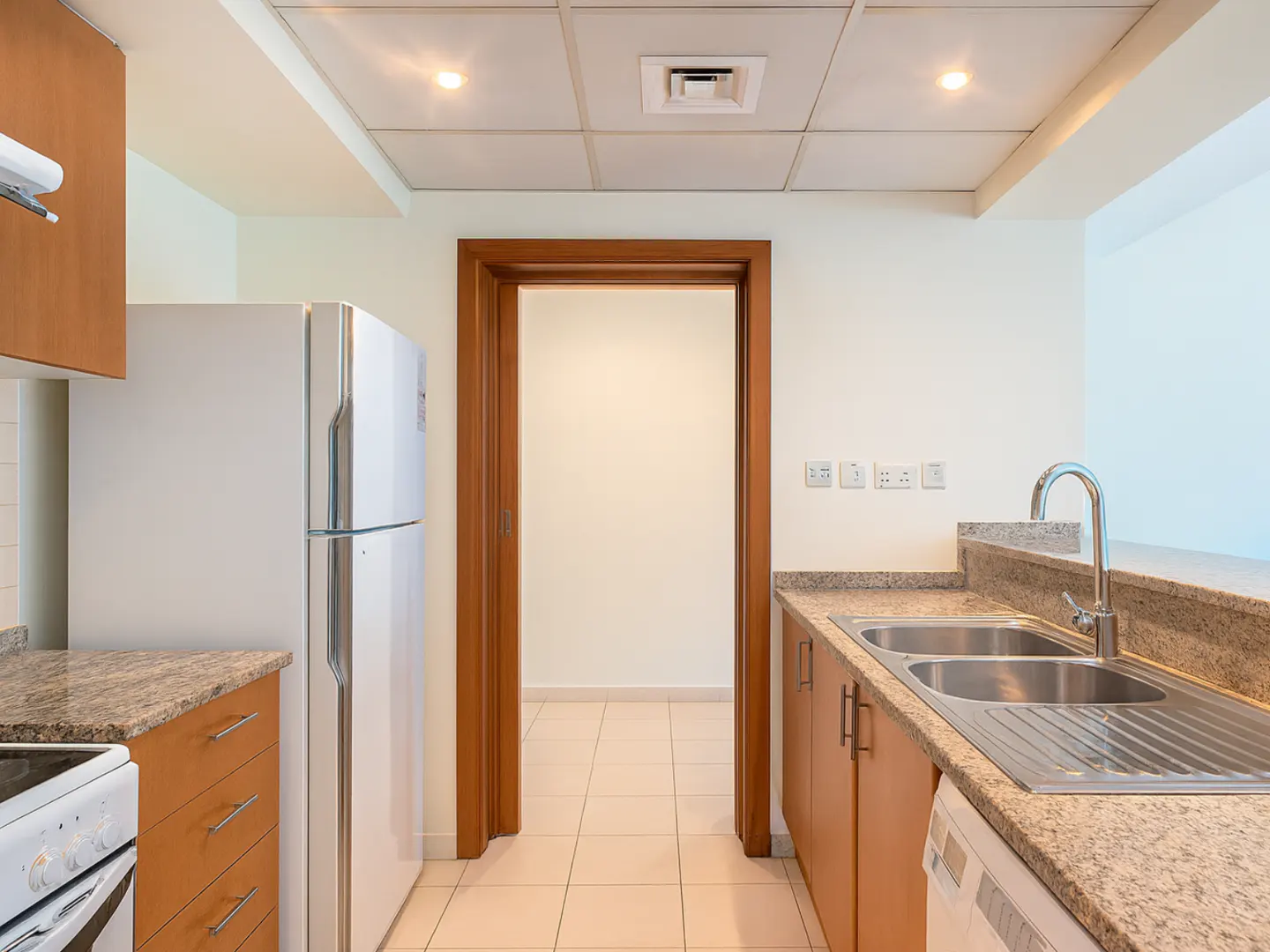 A kitchen with a white refrigerator, brown cabinets, granite countertops, and a stainless steel sink. A doorway leads to another room.