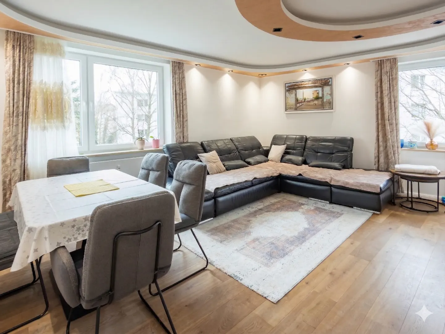 Living room with a black leather sectional sofa, dining table with gray chairs, and hardwood floors. Windows provide natural light.