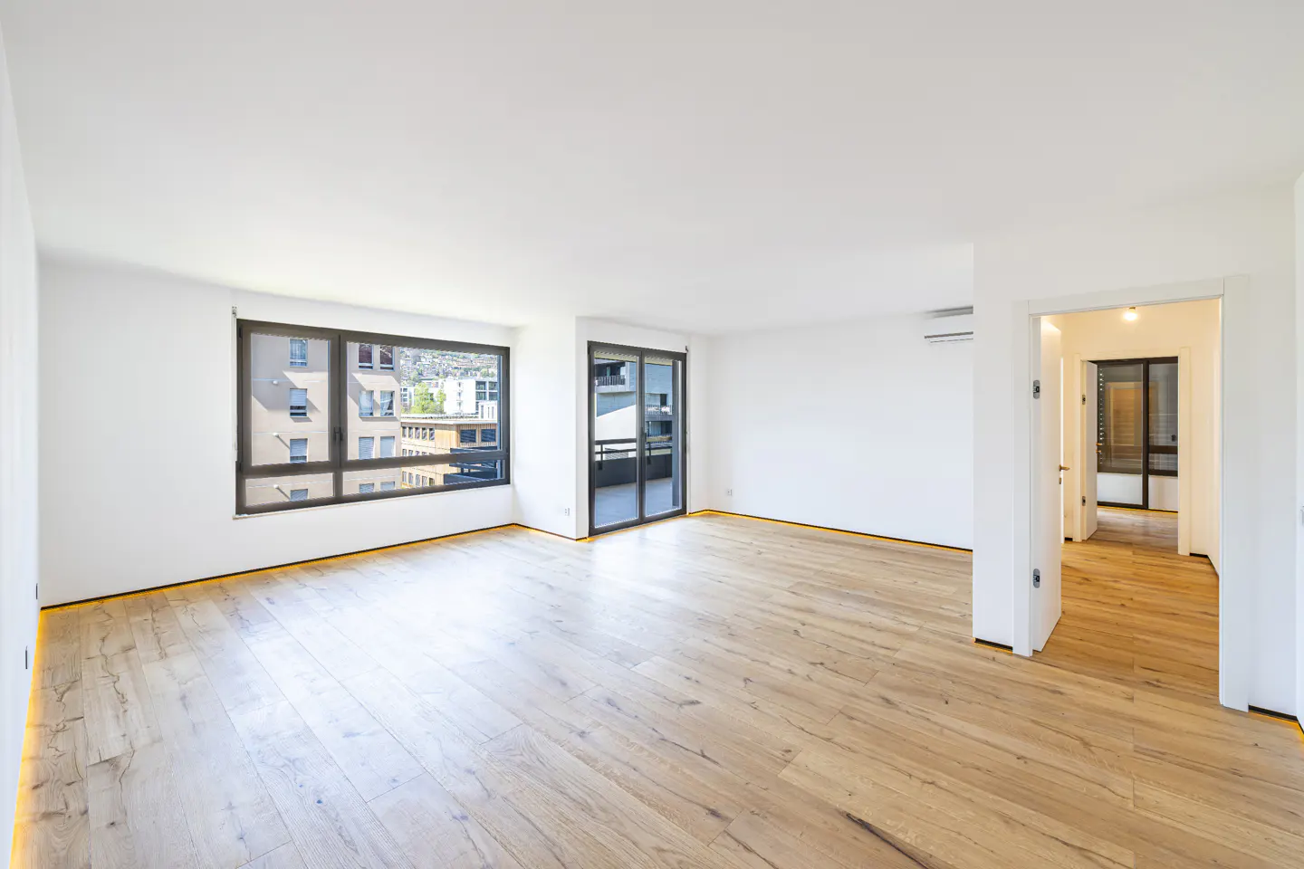 Bright, empty room with light wood floors, white walls, and large windows. A balcony door and open doorway lead to other rooms.