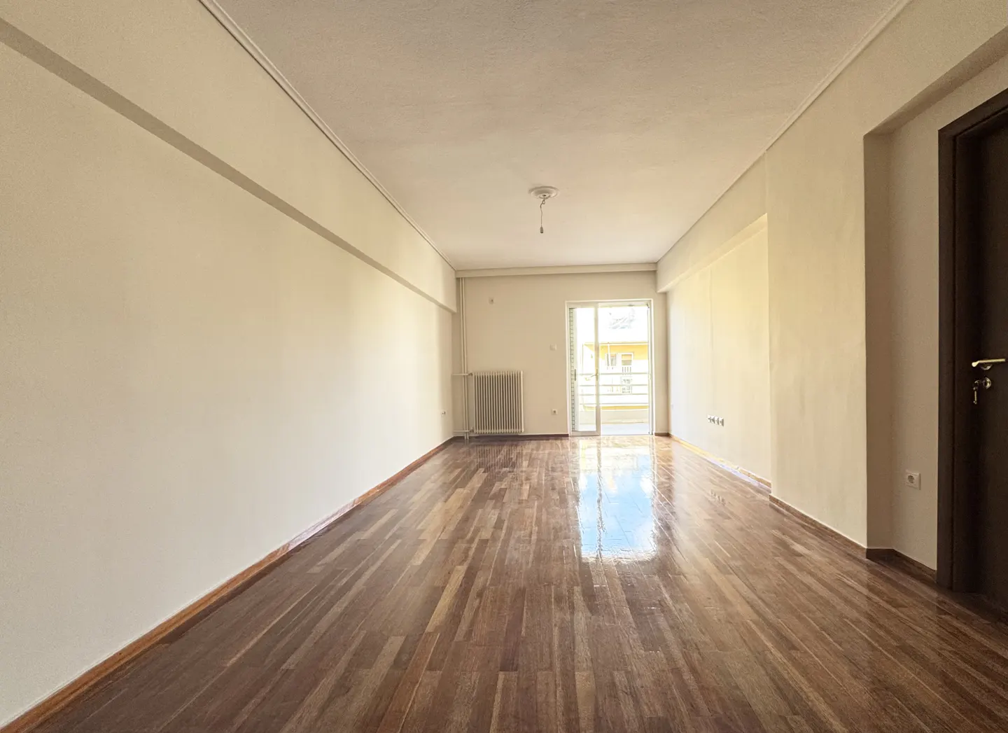 Bright, empty room with shiny hardwood floors, white walls, and a sliding glass door to a balcony. A dark wood door is on the right.