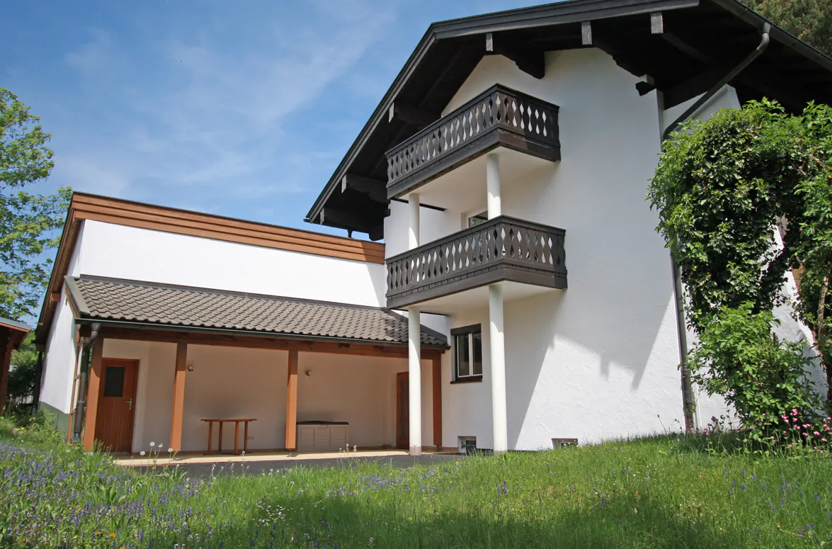 Exterior view of a white two-story house with dark brown trim and two balconies, surrounded by green grass and blue flowers.