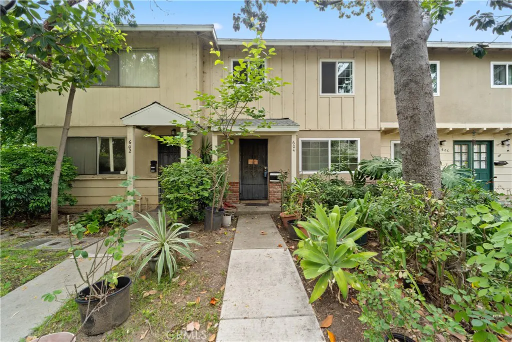 Tan townhouse with a concrete walkway leading to the front door, surrounded by lush green plants and trees.
