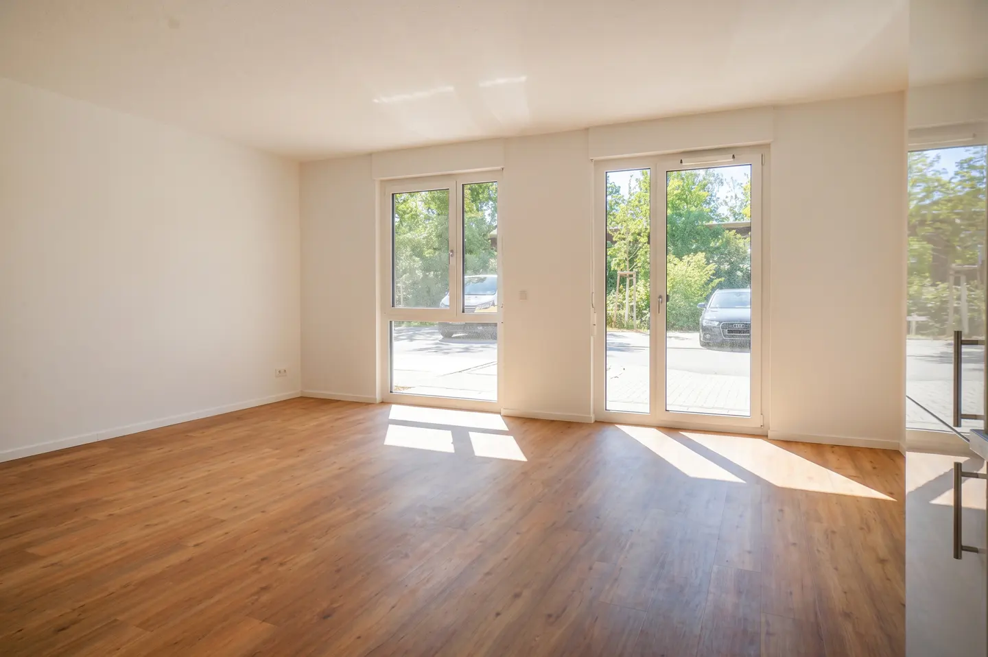 Bright, empty room with white walls and wood floors. Sunlight streams in through glass doors to a green outdoor view.