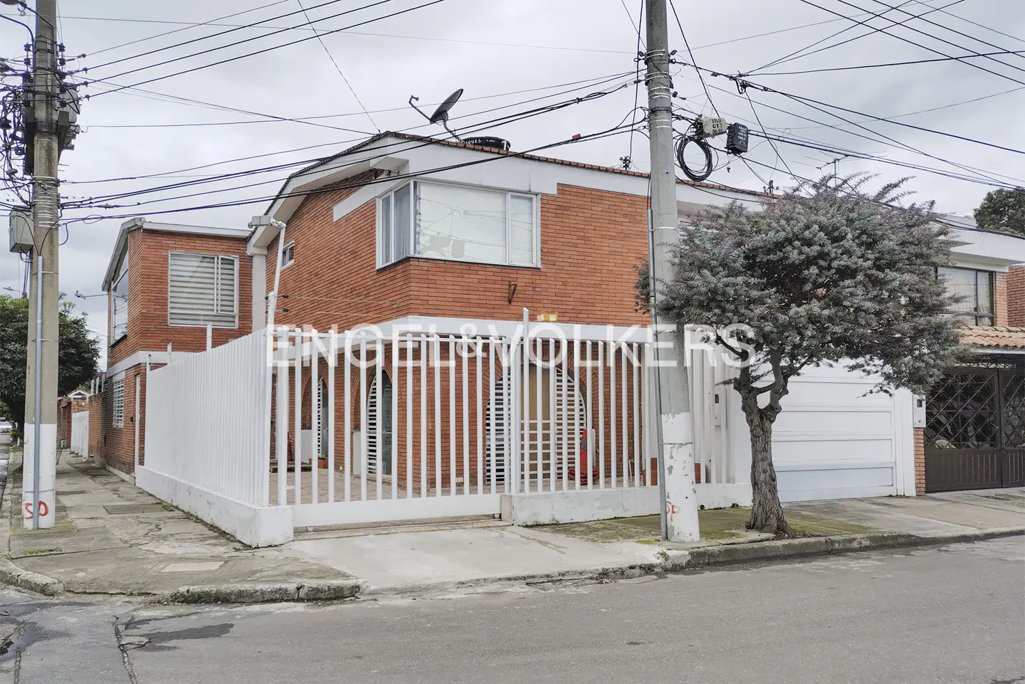 Two-story brick house with white trim and a white fence, viewed from the street on an overcast day.