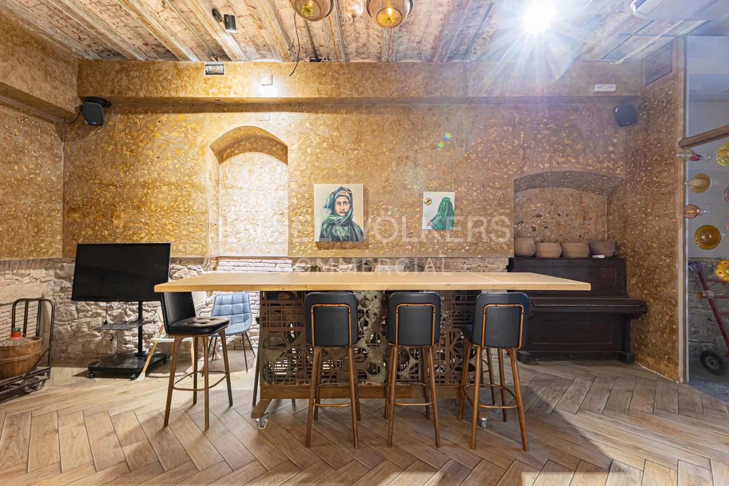 Interior view of a room with a long wooden table and black stools, stone walls, and herringbone wood floors.