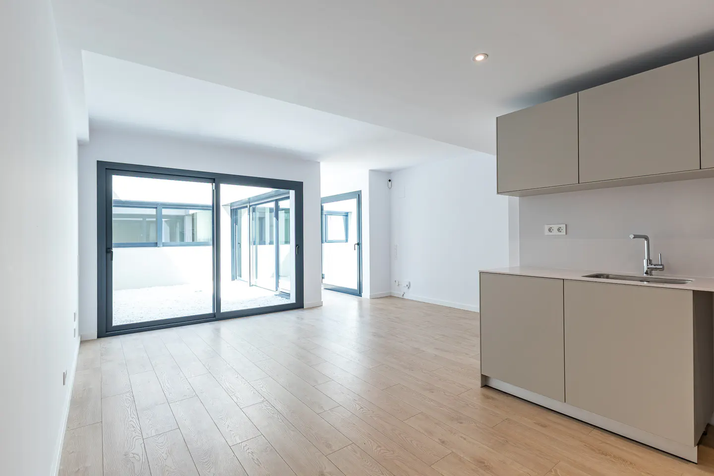 Bright, modern apartment interior with light wood floors, white walls, and gray kitchen cabinets. Sliding glass doors lead to a patio.