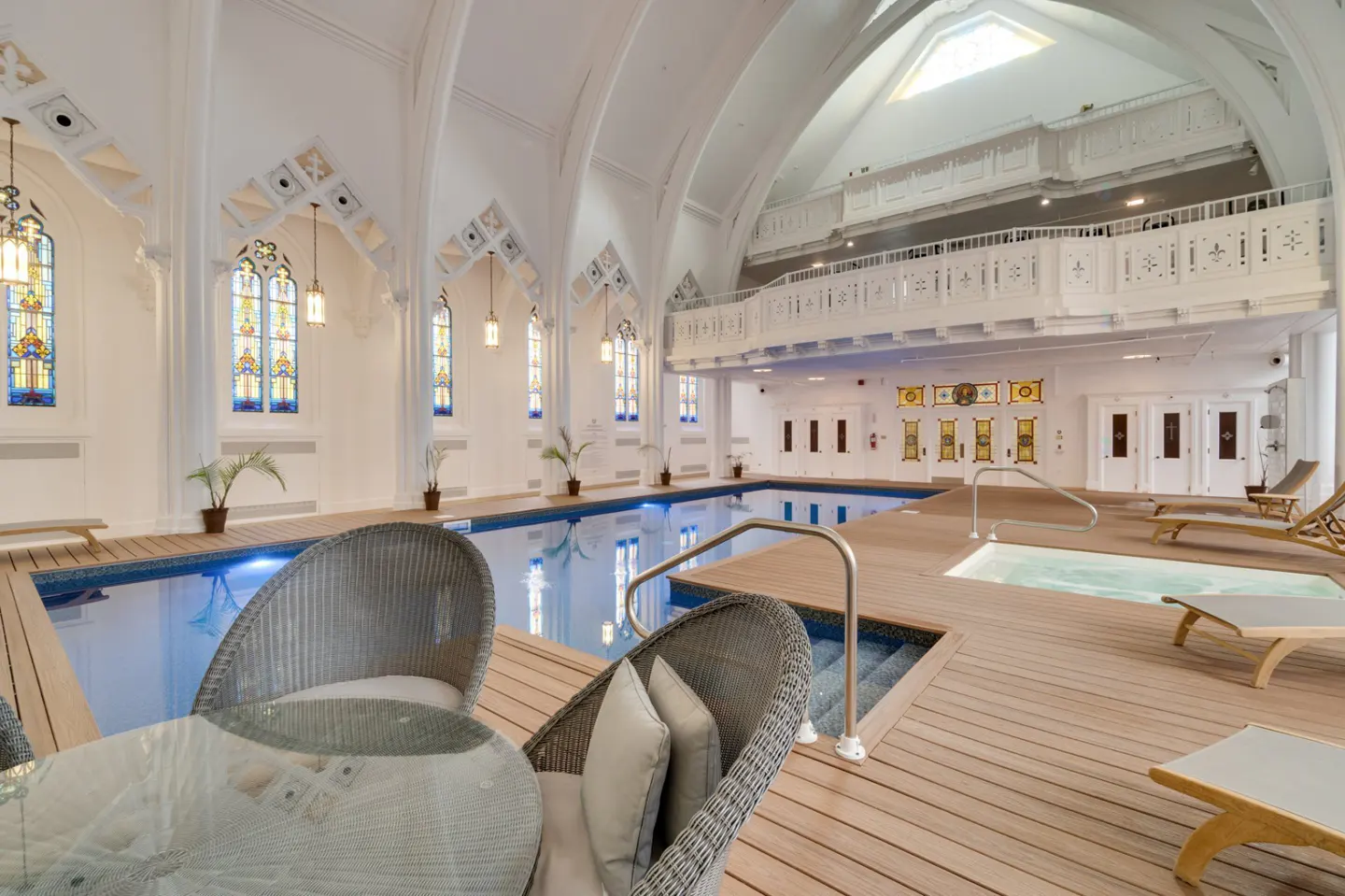 Indoor pool in a converted church with stained glass windows and a balcony. Wicker chairs and a glass table are in the foreground.