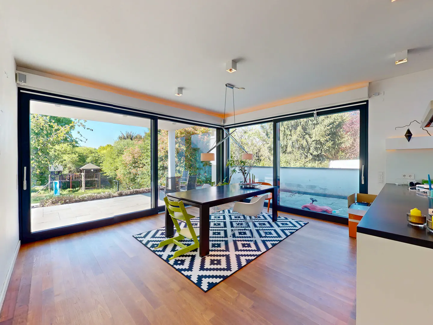 Bright dining area with wood floors, black table, and colorful chairs on a black and white patterned rug. Large windows overlook a green yard.