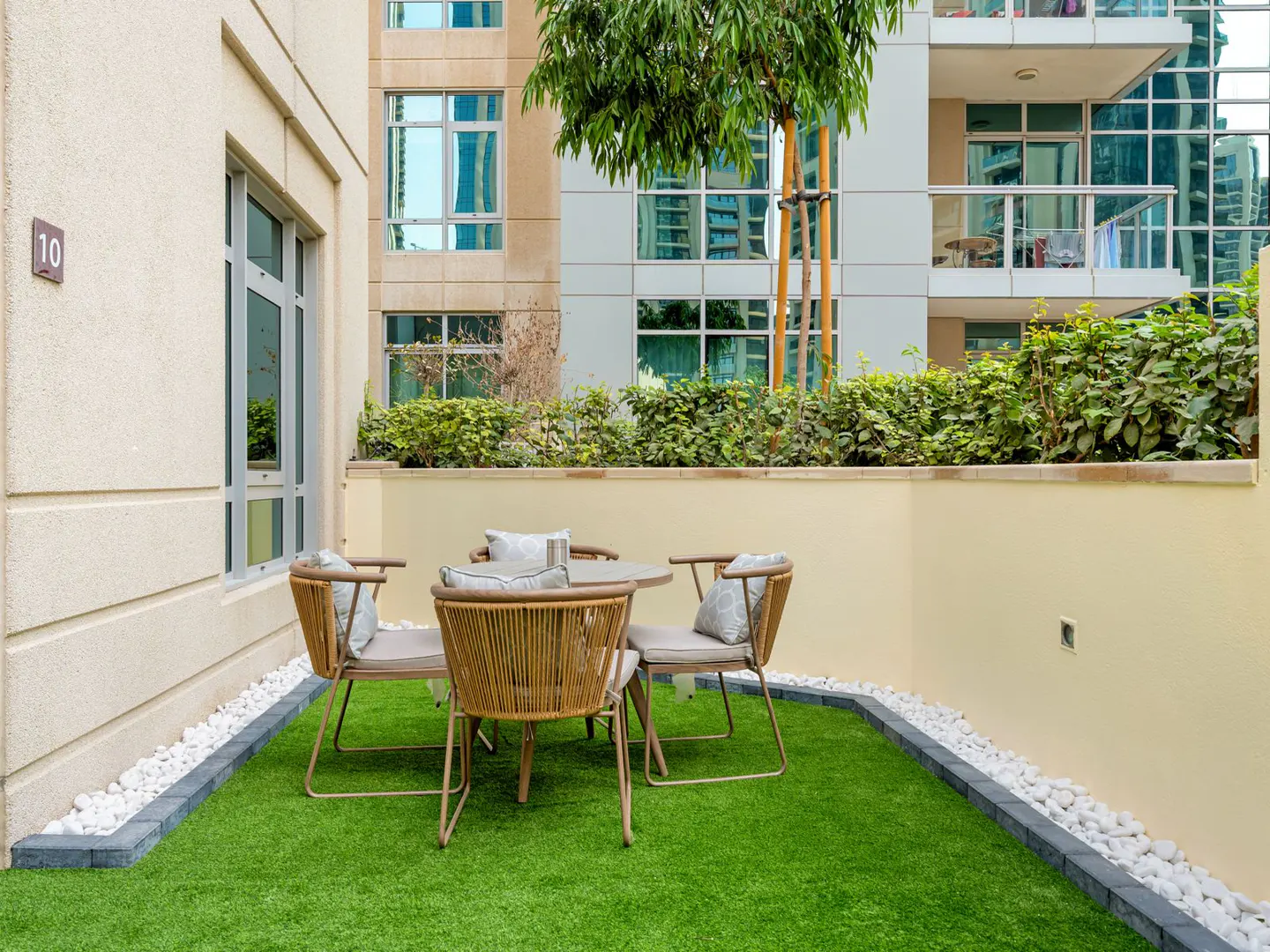 Outdoor patio with four wicker chairs and a round table on green turf. Buildings and greenery in the background.