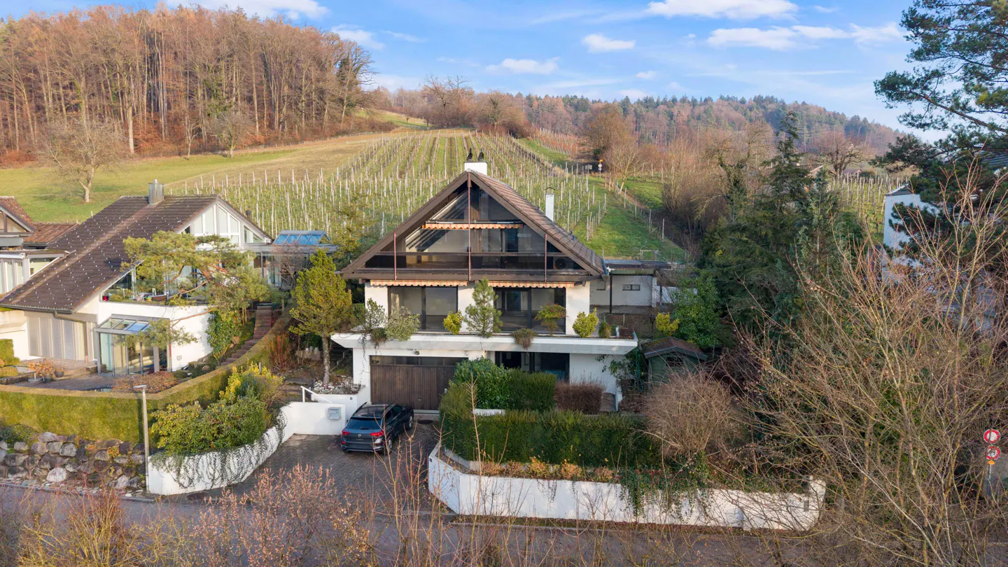 A-frame house with a brown roof and white exterior, surrounded by greenery and a vineyard in the background.