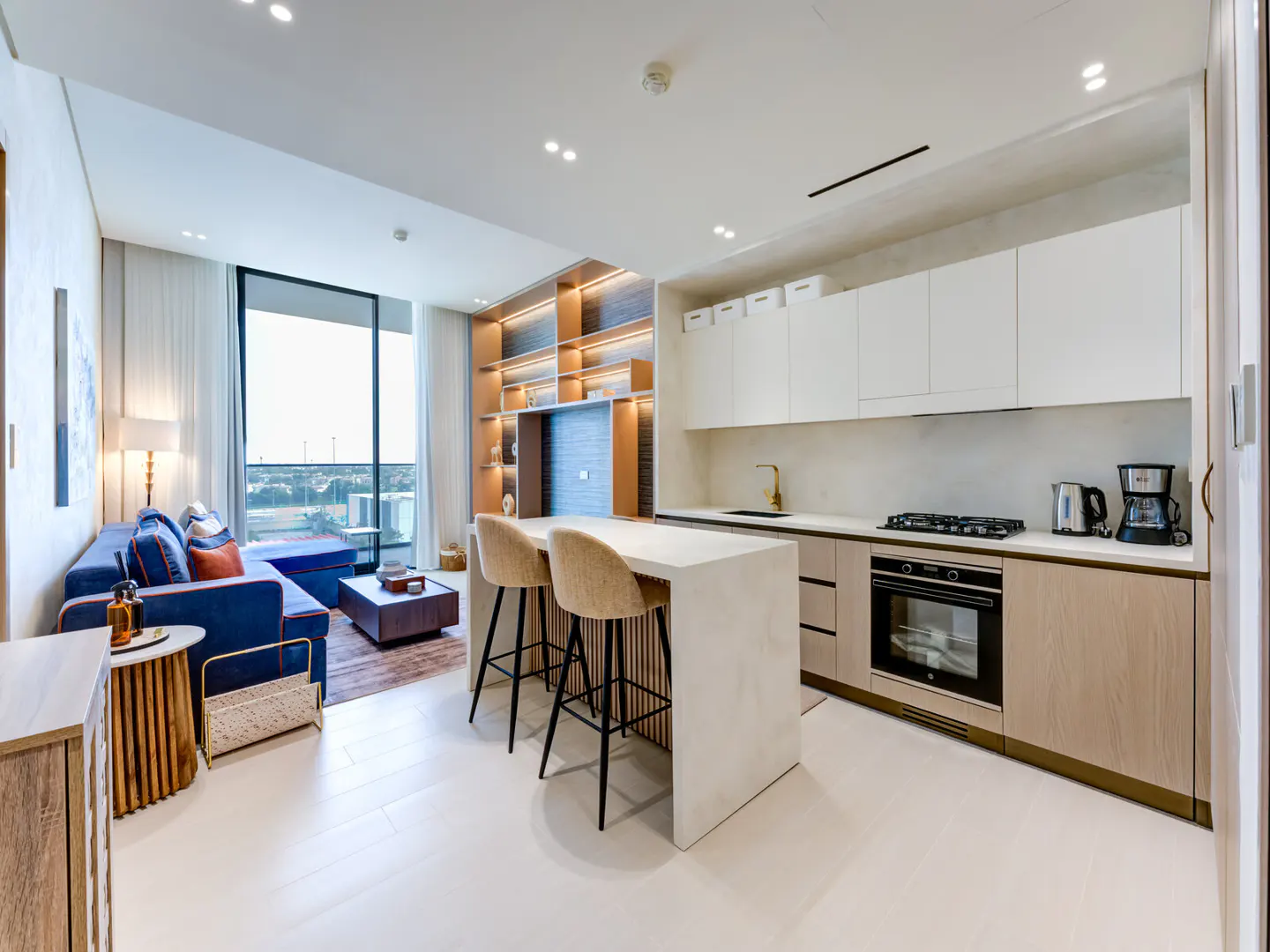 Bright, modern apartment interior with a blue sofa, kitchen island with two stools, and built-in shelving.