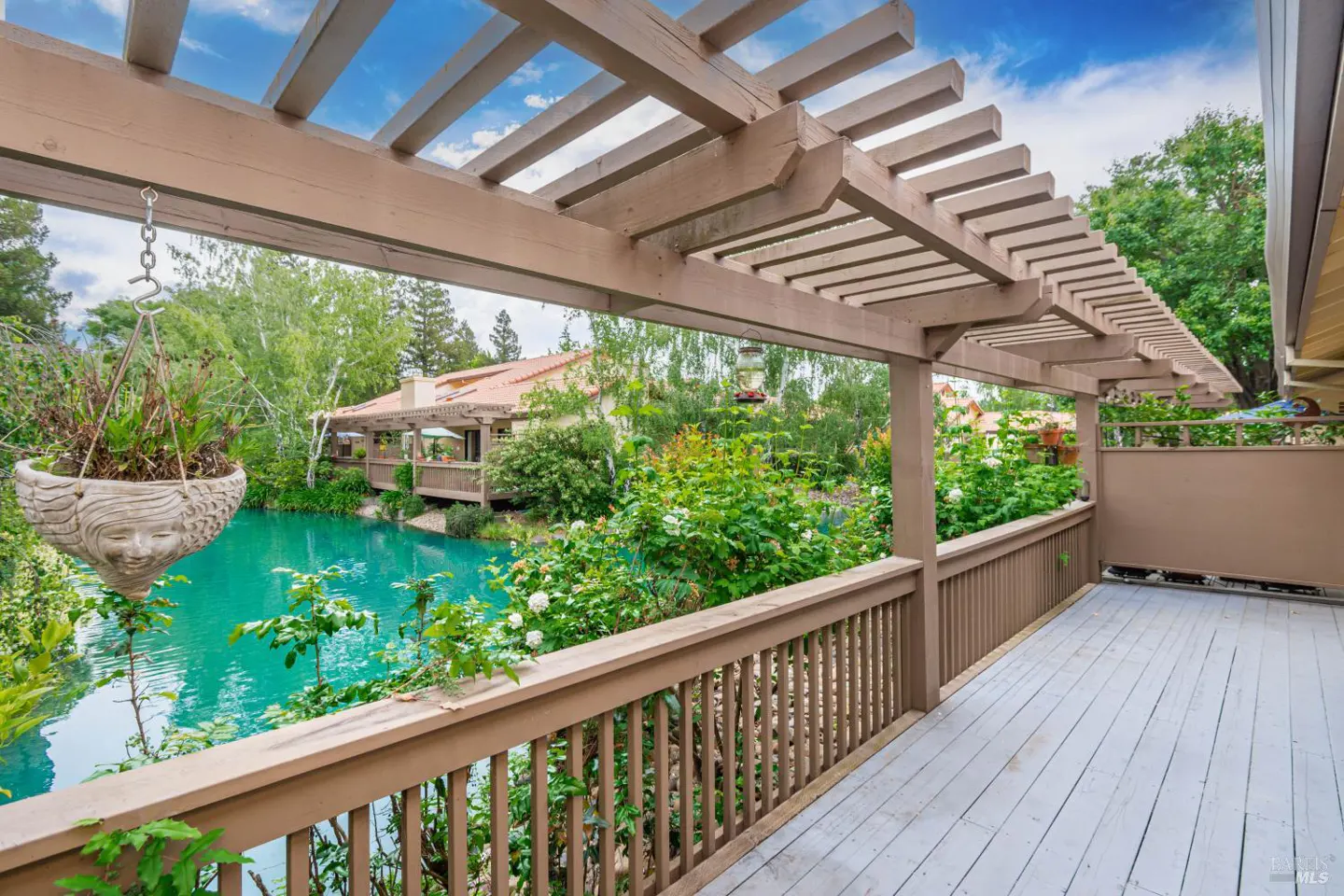 Wooden deck with pergola overlooking a turquoise pond. A hanging planter with a face design is visible. Lush greenery and a building are in the background.